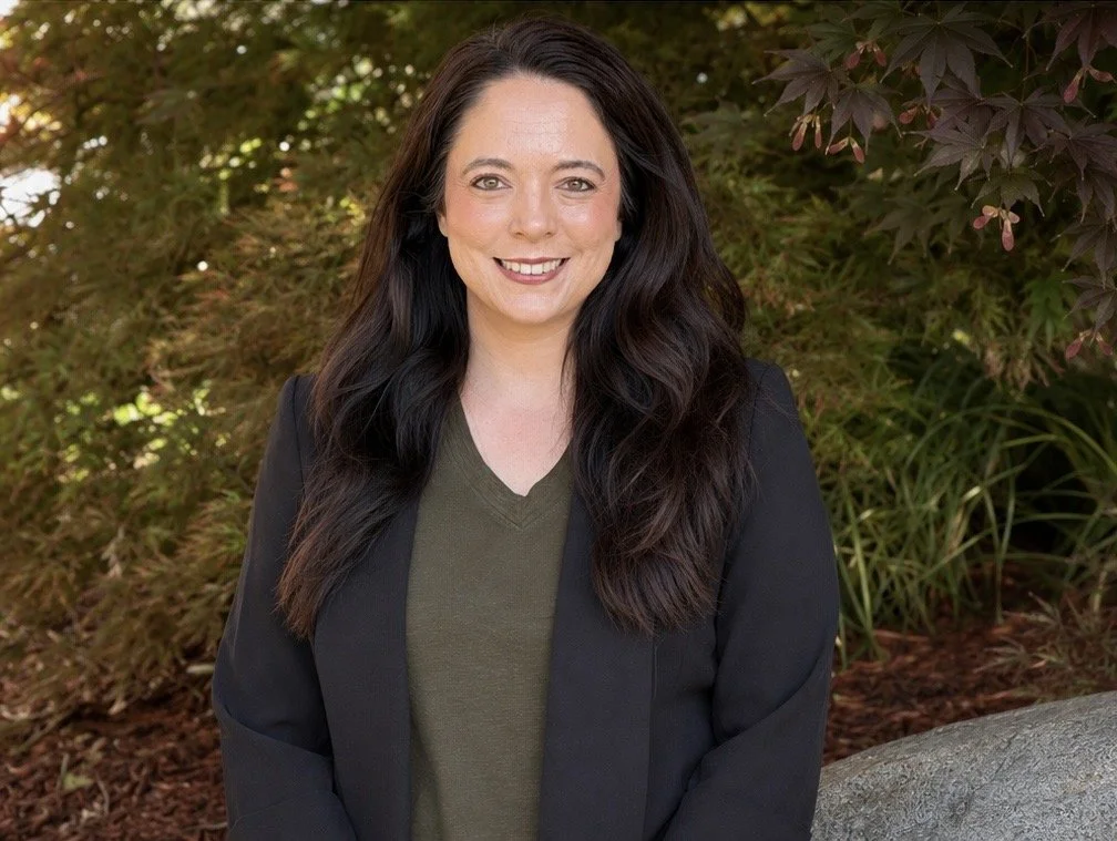 A woman with long dark hair smiling outdoors, wearing a black blazer over an olive green top, with greenery and foliage in the background.