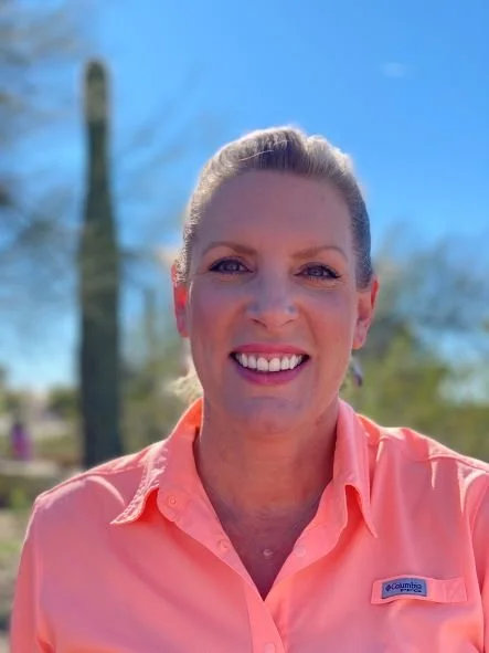 A woman with blonde hair tied back, smiling, outdoors on a sunny day, wearing a peach-colored collared shirt.