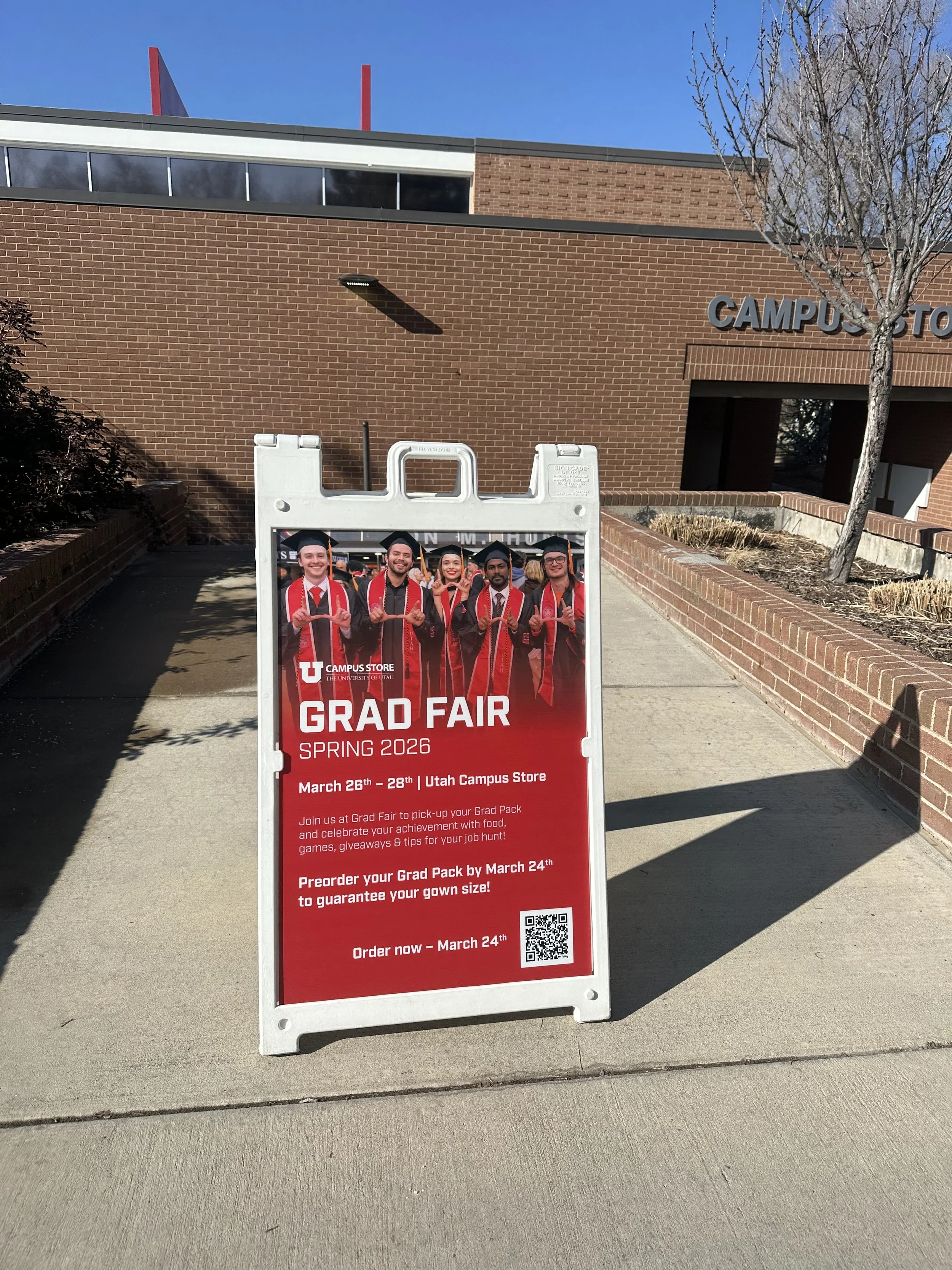 A sidewalk sign advertising a graduation fair at the University of Utah campus store, happening from March 26 to 28, Spring 2026.