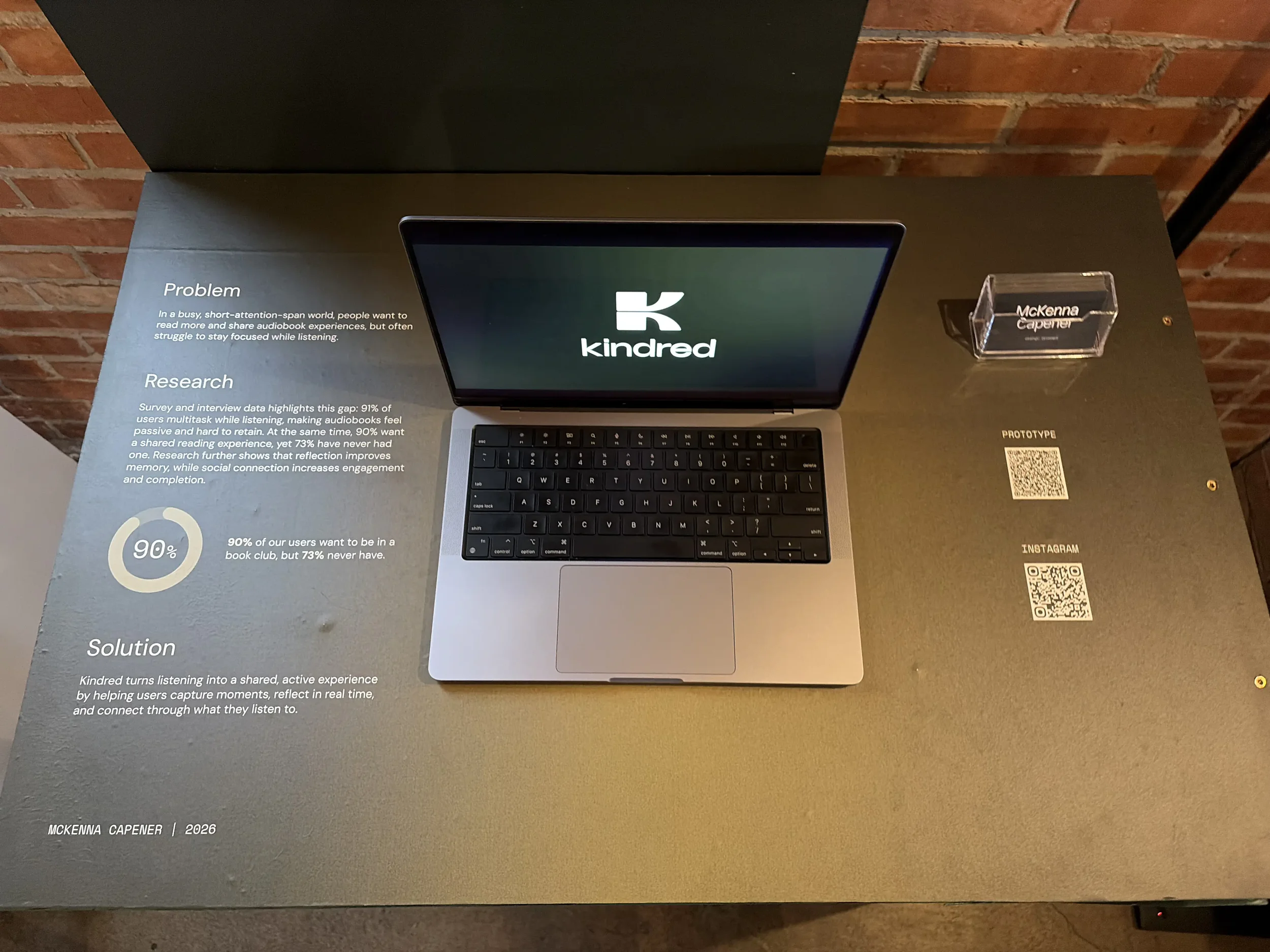 Exhibit display featuring a MacBook laptop with the Kindred logo on its screen, a small clear nameplate reading 'McKenna Capener,' and informational text about Kindred's mission to enhance audiobook experiences, with a brick wall in the background.