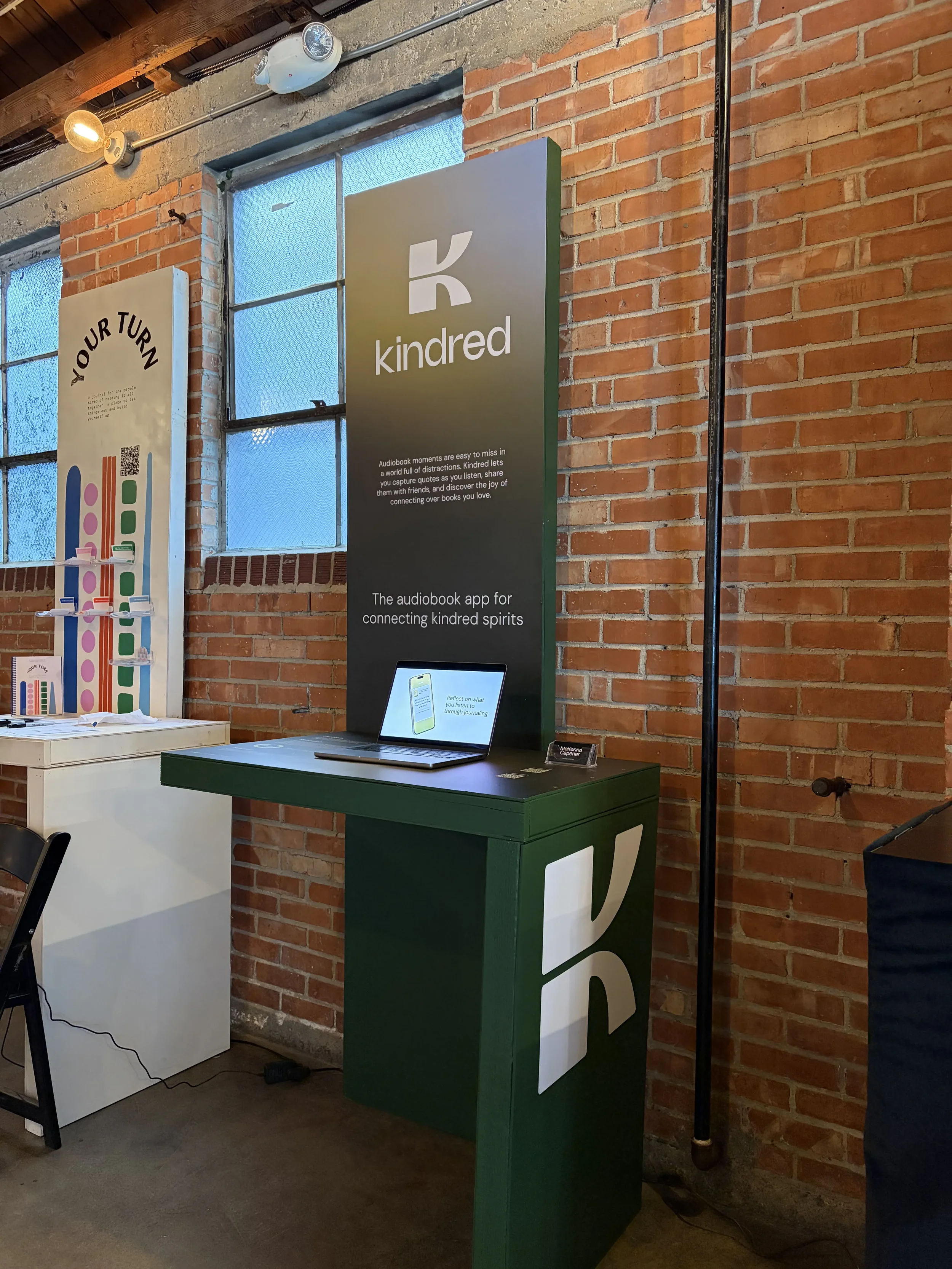 Inside a brick-walled space with a window, there is a green and white kiosk labeled 'kindred' and a laptop on top of it. To the left, a white display with colored abstract shapes stands next to a white table. The space has exposed wooden ceiling beams and lighting fixtures.
