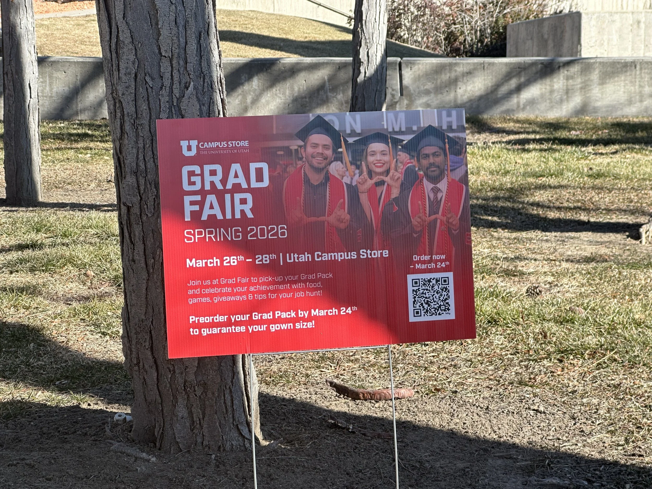 Red poster on a wooden tree stand announcing a graduation fair at Utah Campus Store with dates March 26th to 28th, 2026, featuring photos of three people in graduation caps and gowns.