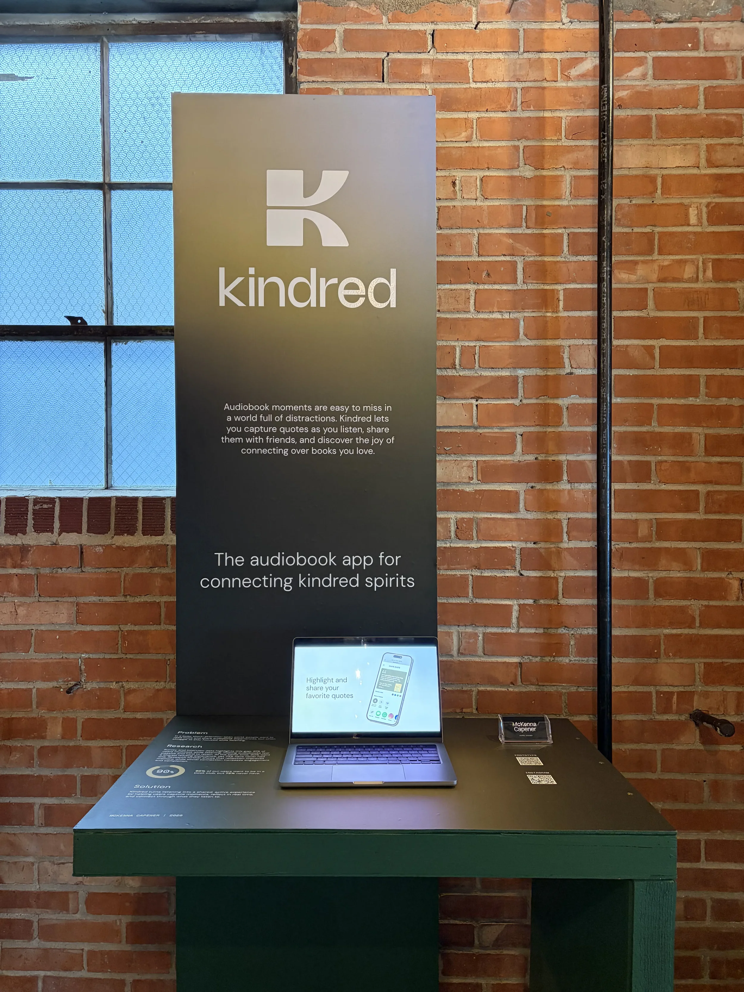 A display stand for Kindred, an audiobook app, featuring a sign with the Kindred logo, a laptop showing the app interface, and information about capturing quotes, set against a brick wall.
