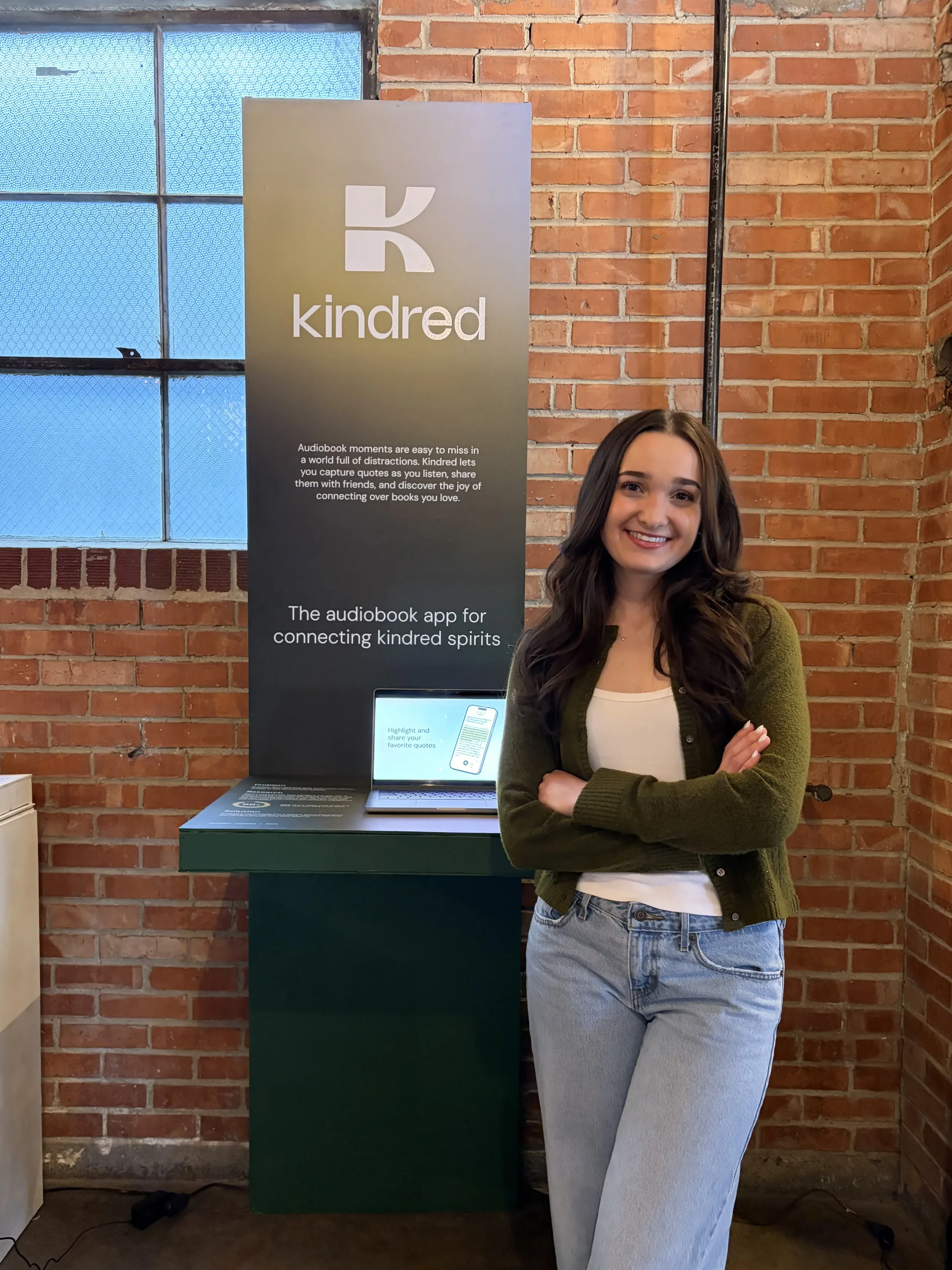 A young woman with long brown hair, wearing a green cardigan and light blue jeans, is standing with her arms crossed and smiling in front of a promotional display for Kindred, an audiobook app, in an indoor space with exposed brick walls and a window with textured blue glass.