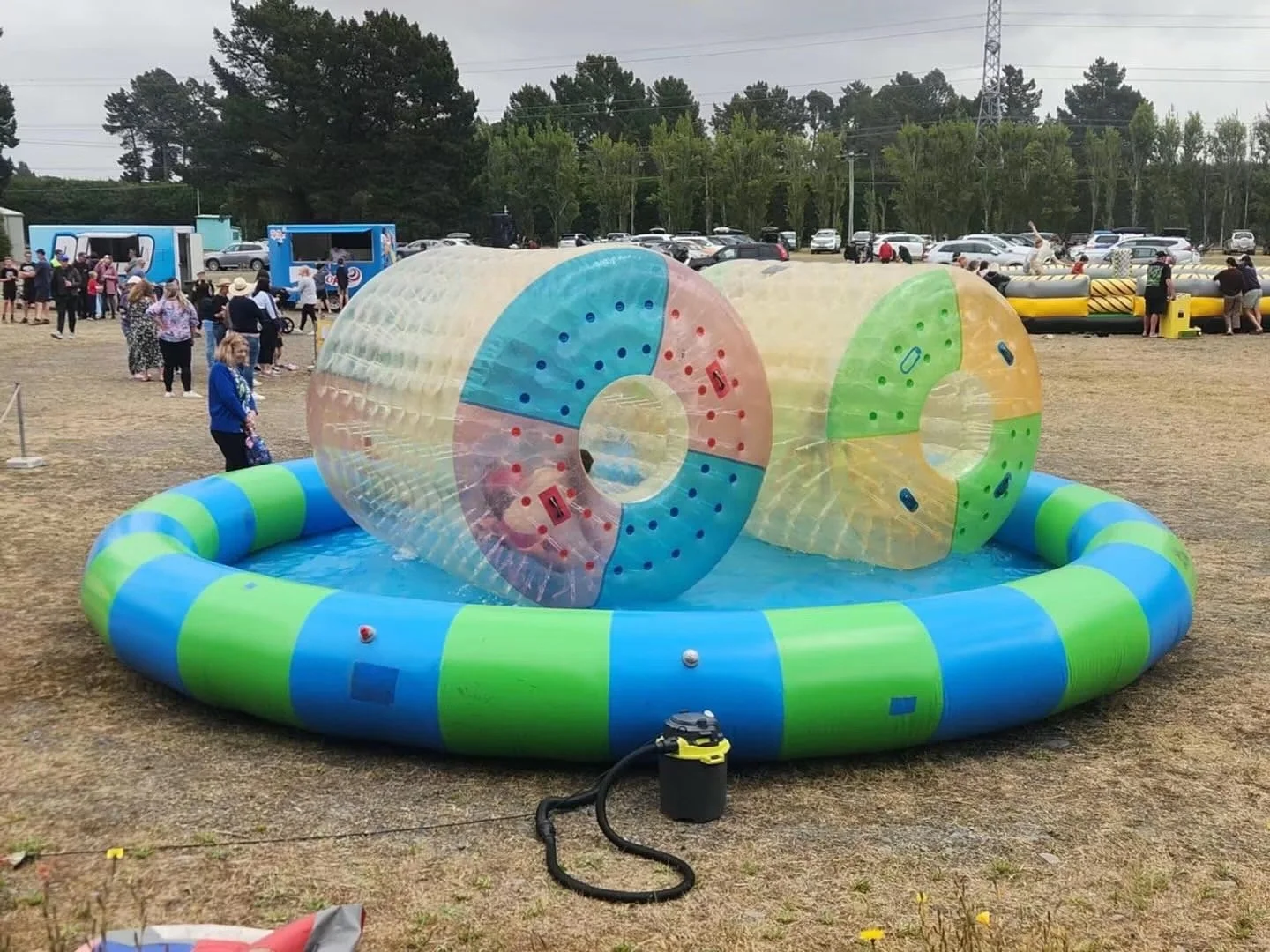 Inflatable water obstacle course with large transparent plastic spheres on a grassy field, surrounded by people, trucks, and trees in the background.