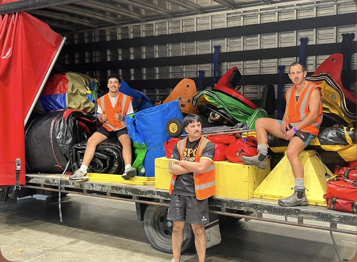 Three young men in orange safety vests posed on a vehicle loaded with bags, equipment, and trash, inside a storage or garage facility.
