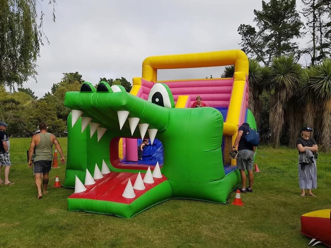 Colorful inflatable obstacle course resembling a crocodile with sharp teeth, set up outdoors on grassy area with trees in background, surrounded by people.