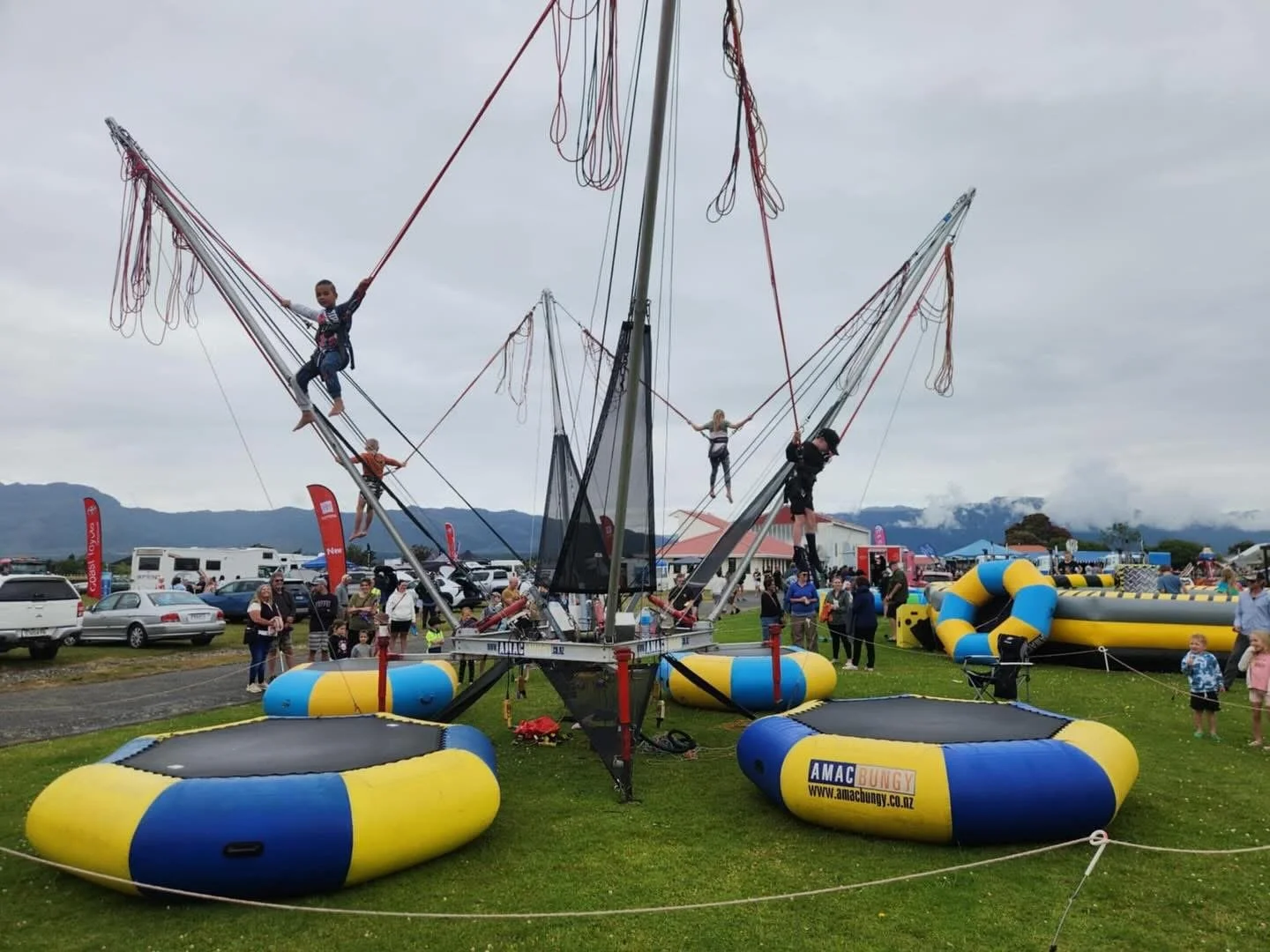People at an outdoor event climbing and playing on an inflatable trapeze ride with bouncy trampolines on the ground.