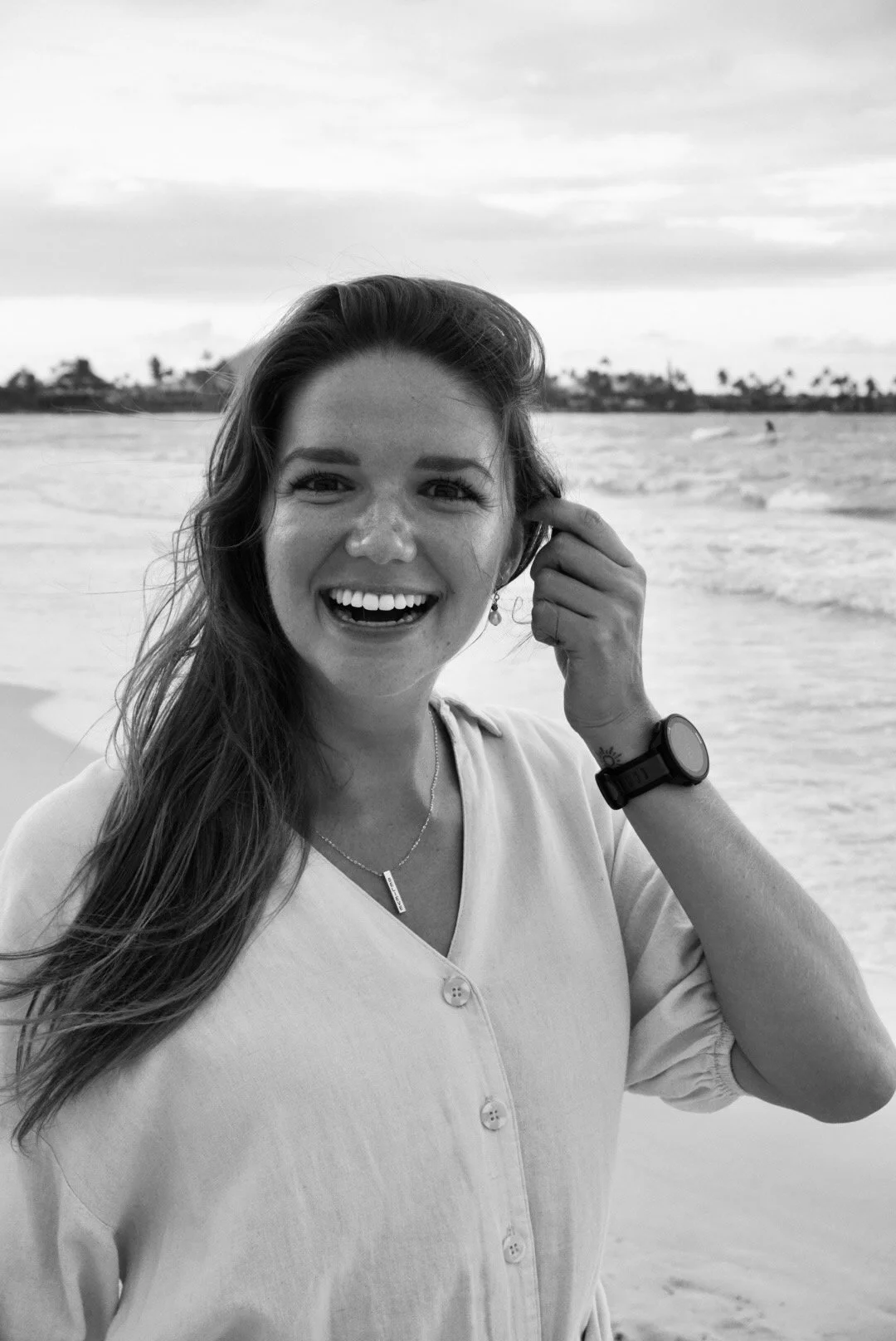 Vineta Gleba, counselor and coach. Black and white photo of a kind, smiling woman at the beach, with ocean waves and palm trees in the background. She has long hair, is wearing a button-up shirt, a necklace, earrings, and a watch on her left wrist.