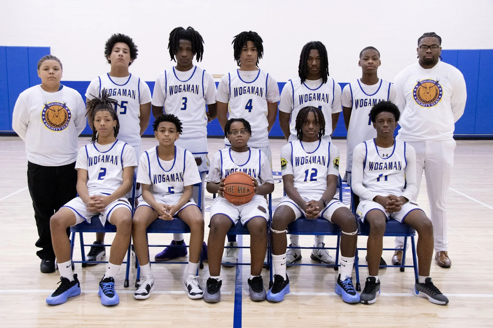 A youth basketball team posing for a group photo in a gymnasium. The team includes children and teenagers in white and blue uniforms, with two coaches or staff members standing on each side. One player in the center is holding a basketball.