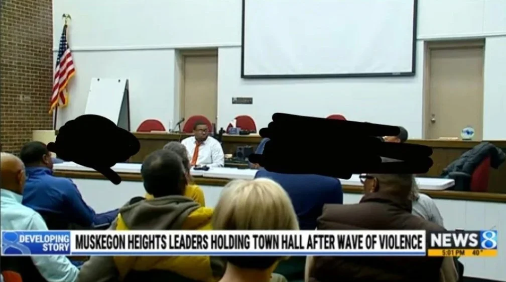 A town hall meeting in Muskegon Heights with several people seated and a speaker at the front, American flag in the background, and a news banner about violence.