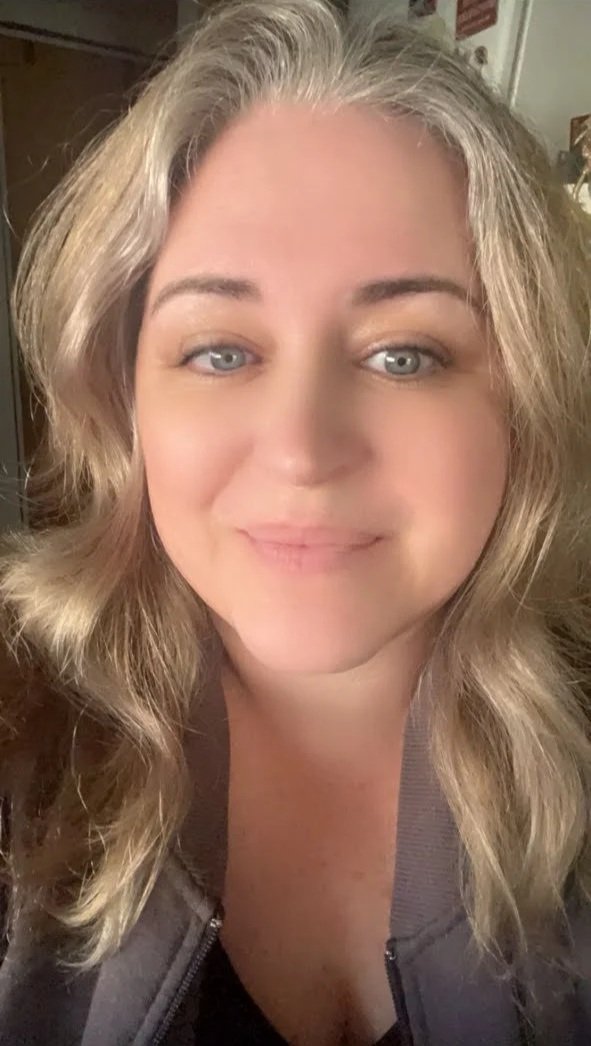 A close-up of a woman with blonde wavy hair, blue eyes, and a light complexion, smiling at the camera.