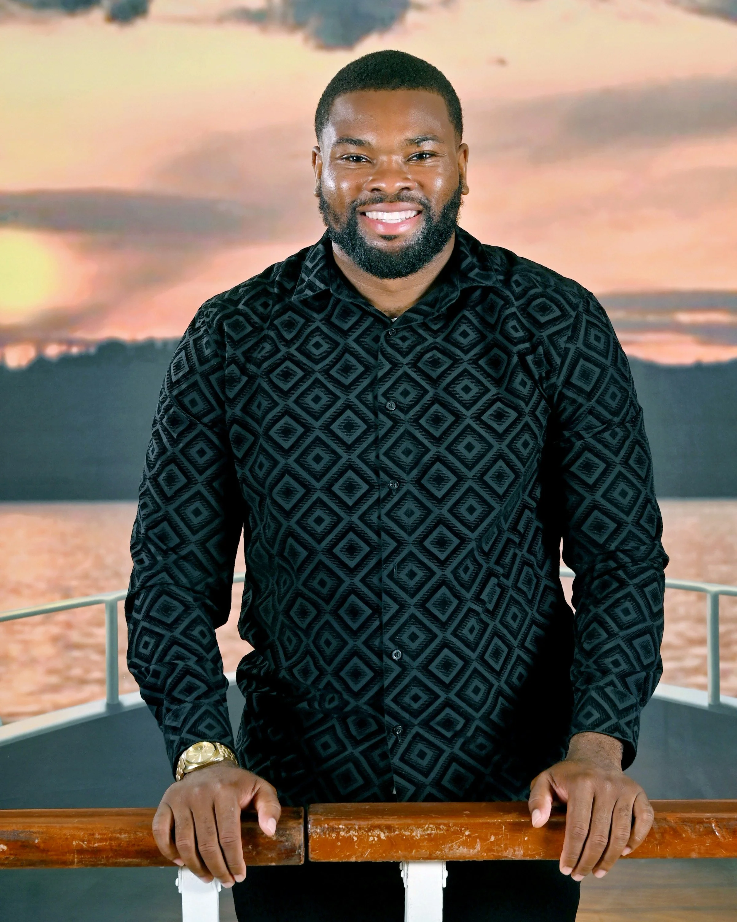 A man with a beard and short hair smiling, standing behind a wooden railing with his hands resting on it. He is dressed in a black geometric patterned shirt and a gold watch. The background features a scenic view of a sunset over the water with clouds in the sky.
