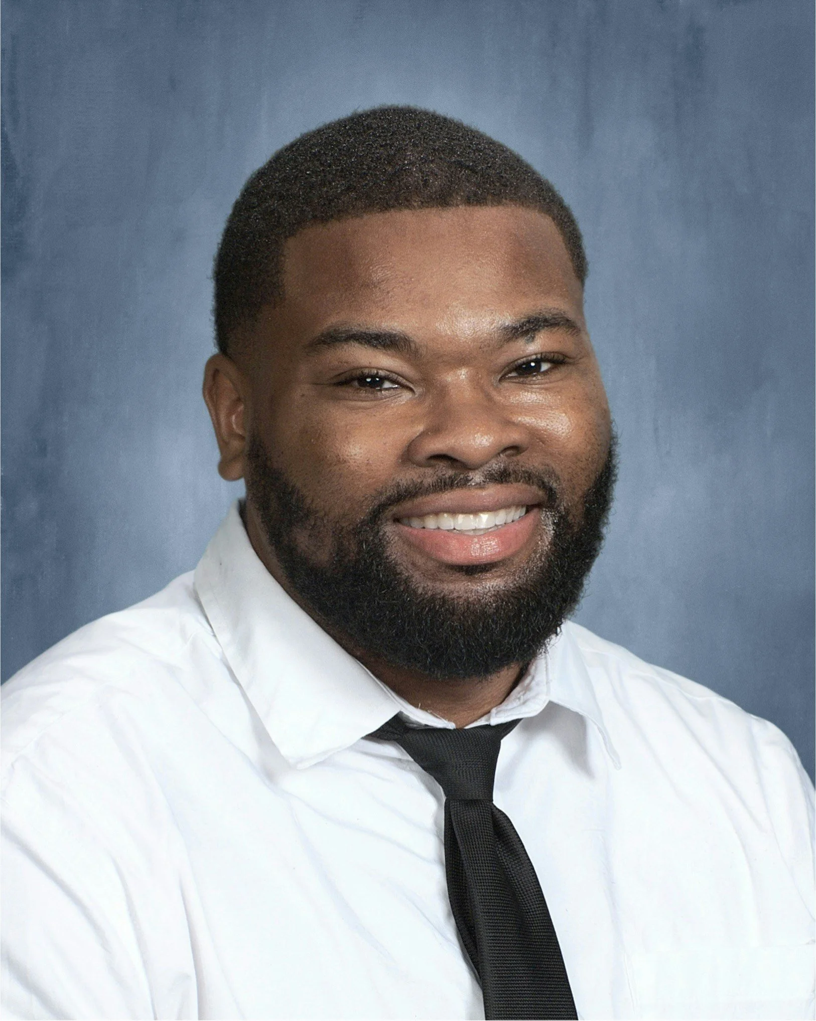 Close-up photo of a smiling man with a beard, wearing a white shirt and a black tie, against a blue background.
