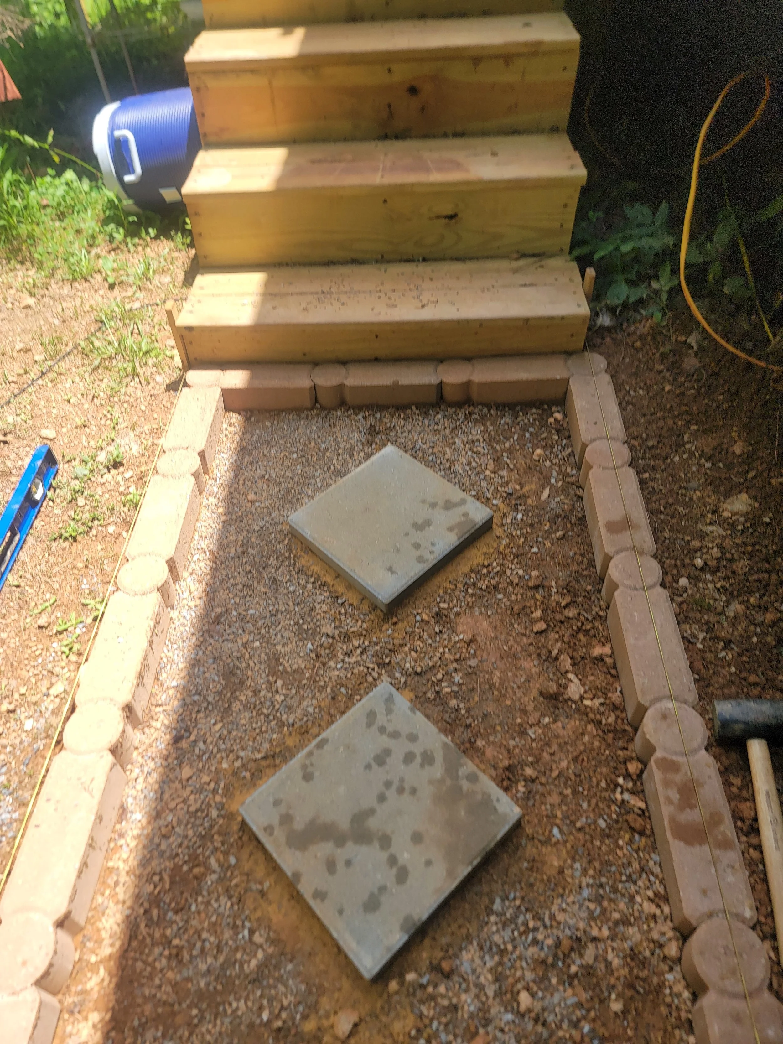 Construction of a wooden staircase with concrete steps, gravel ground, and bricks outlining the stairs, with gardening tools and a blue water container nearby.