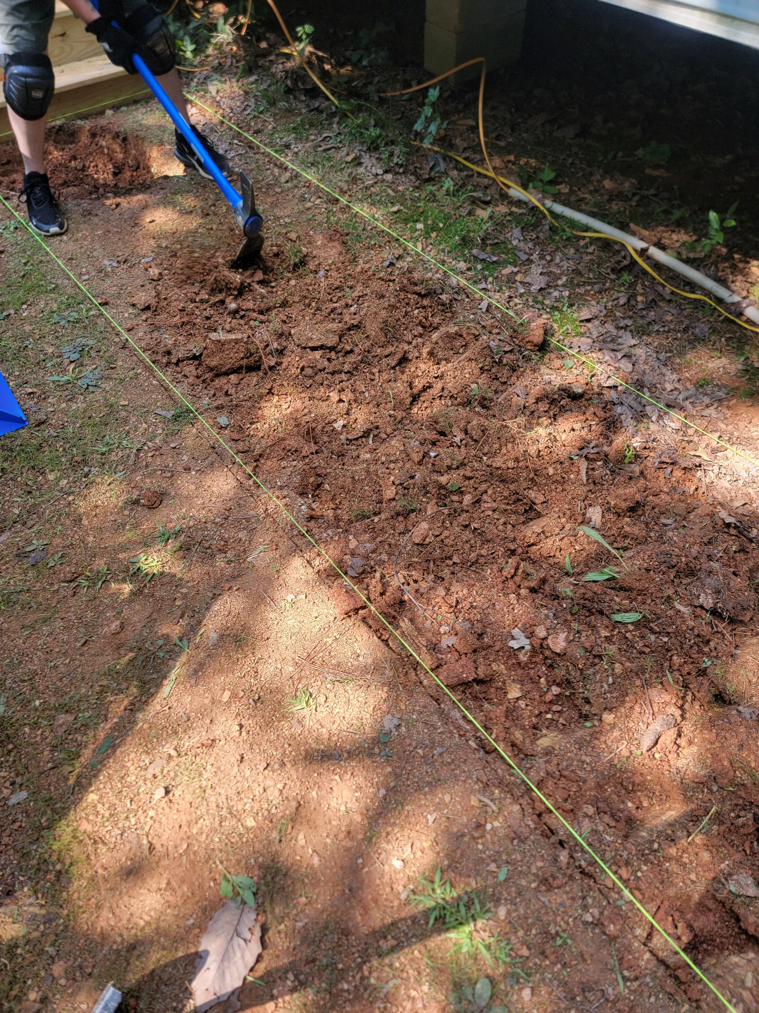 A person is using a small pickaxe to dig a straight trench in the ground, which is marked by yellow string stretched between two stakes. The area has brown soil with some grass and leaves, and there are some yellow and white tubes or pipes along the edge of the digging site.