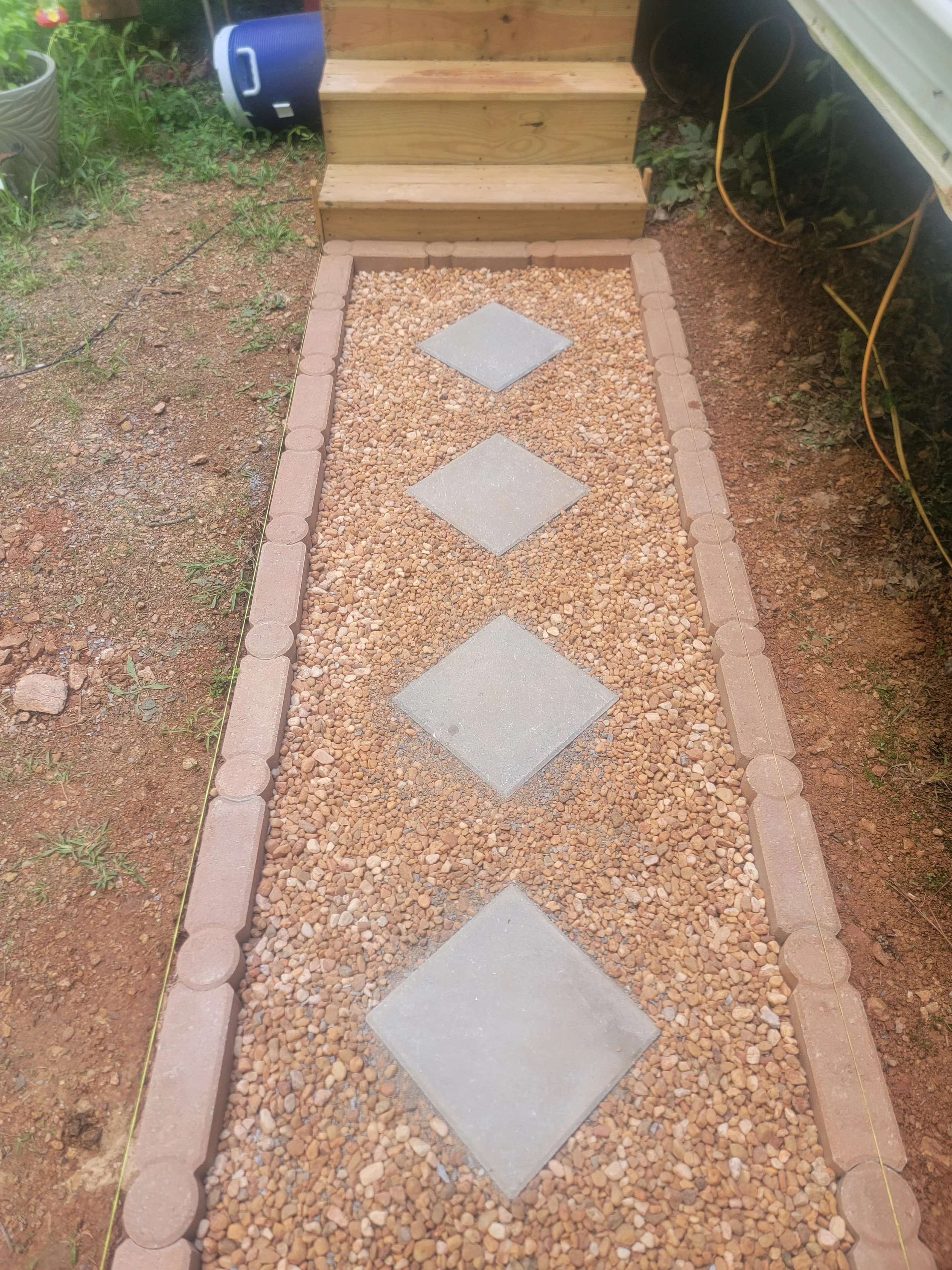A gravel pathway with large square concrete stepping stones, bordered by pinkish brick edging, leading to wooden stairs and a small outdoor space.