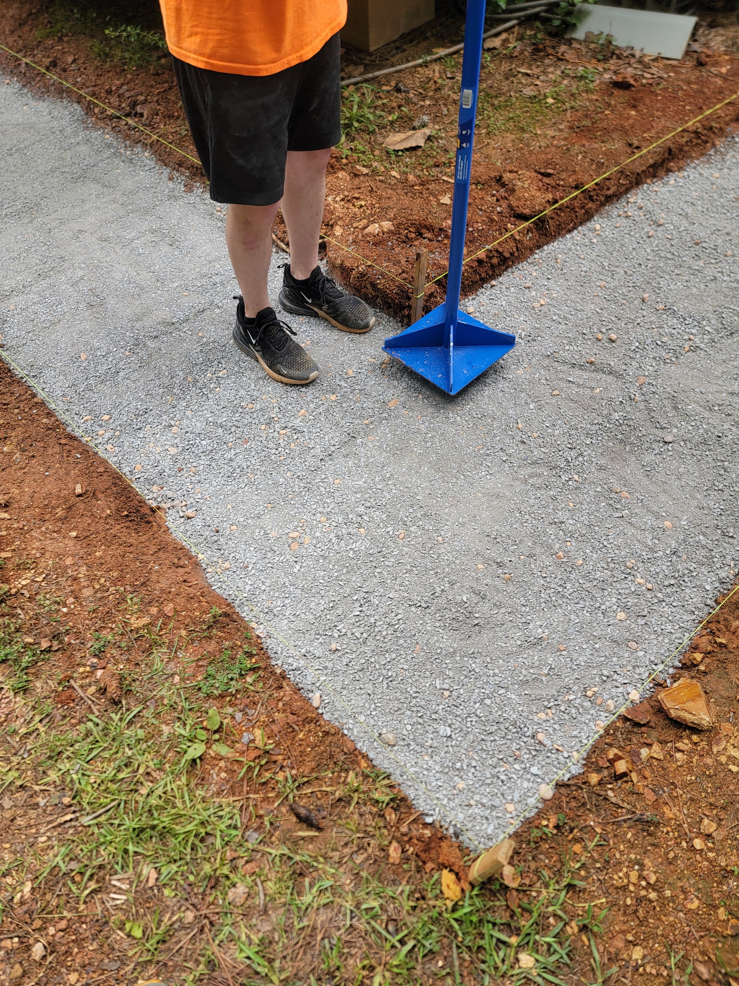 Person in black shorts and black sneakers standing near a freshly laid gravel path with yellow string guides and blue stake, holding an orange shirt, with soil and grass around.