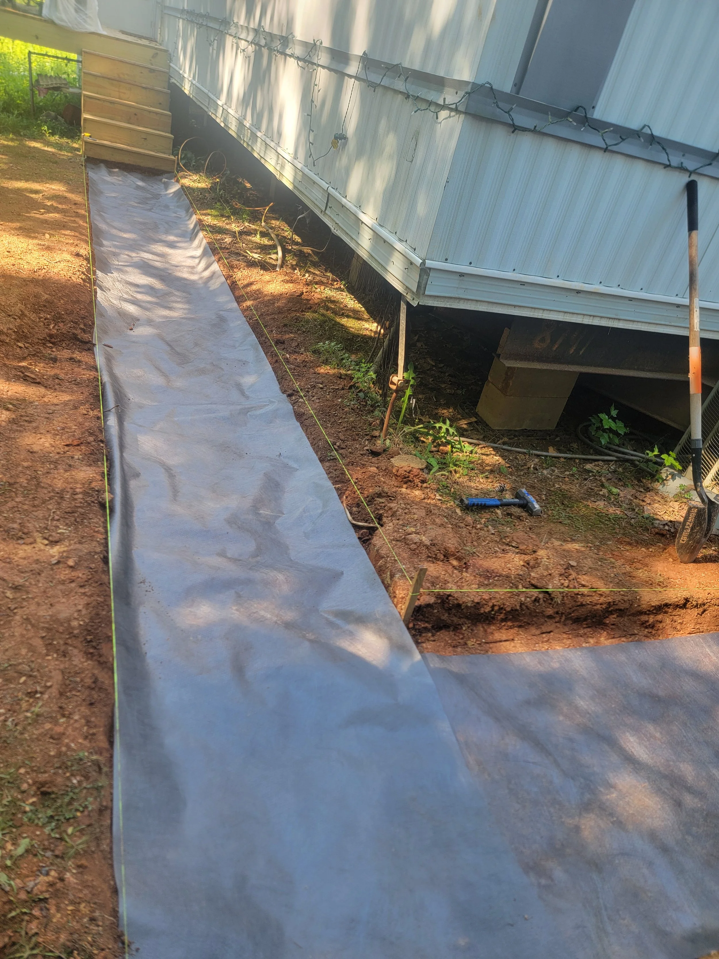 A makeshift walkway with a sheet of clear plastic or tarp on top, set up along a dirt alley next to a raised mobile home. The mobile home has festive string lights attached to it, and there is gardening equipment, including a small shovel and some hoses, visible nearby.