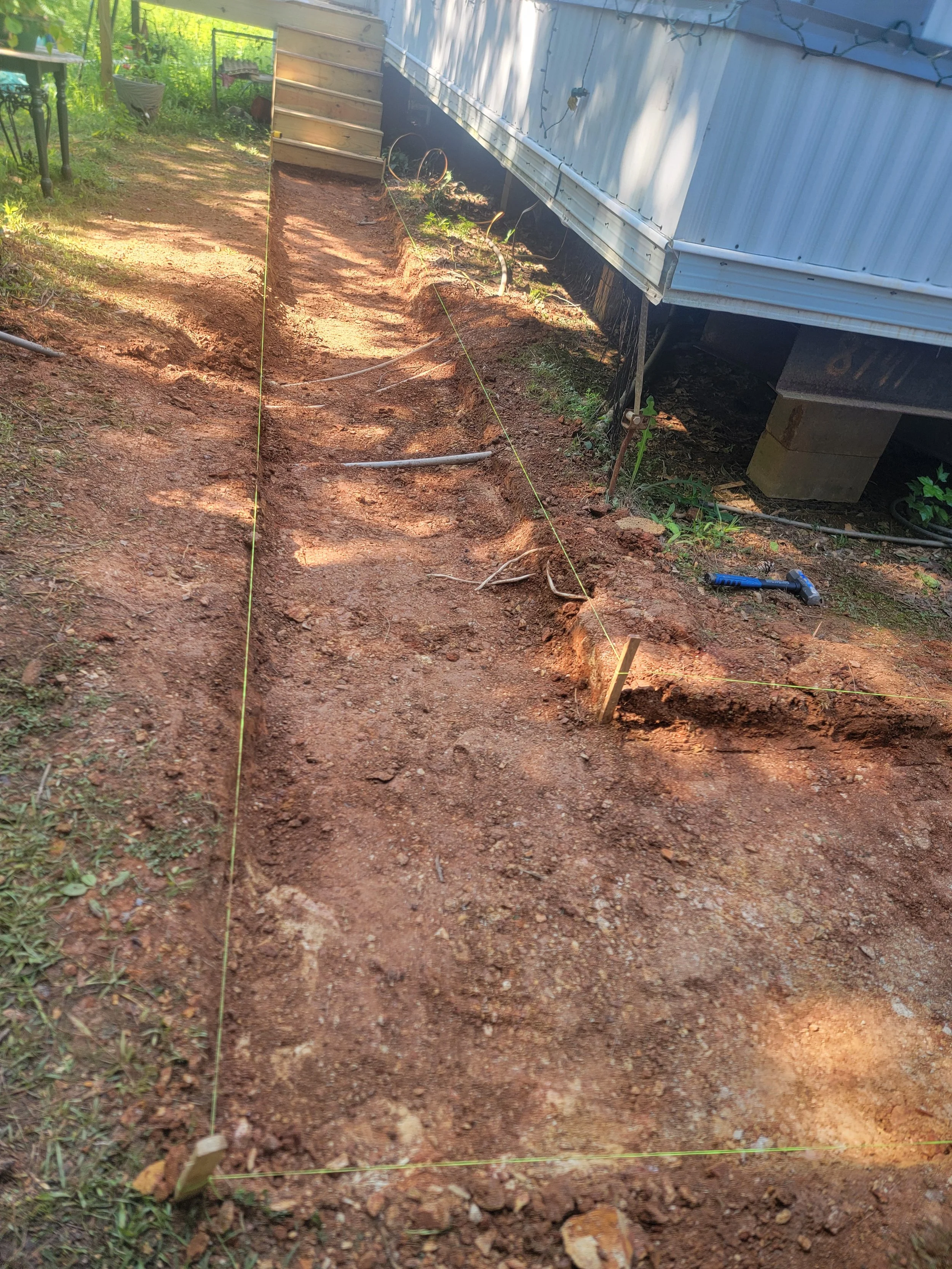 A trench has been dug in a backyard yard area, marked with yellow string for alignment. There are various tools and materials nearby, including a hammer and pipes. The fence and part of a deck are visible in the background.