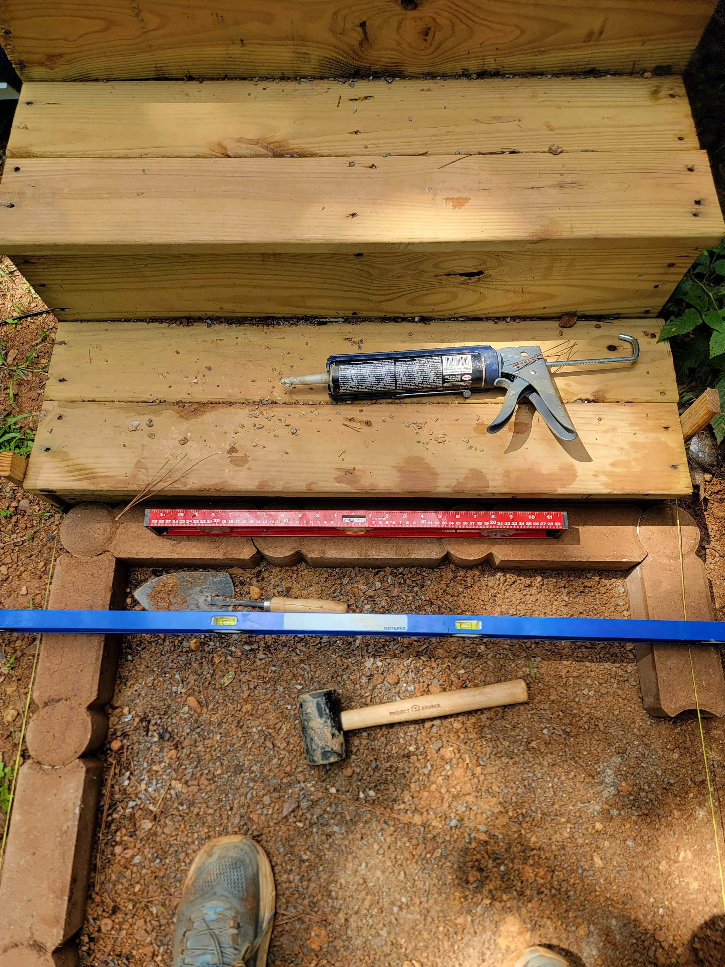 New wooden steps being built with gardening or construction tools such as a caulking gun, impact driver, hammer, and level on the ground in front of the steps, with a person's shoes visible at the bottom of the image.