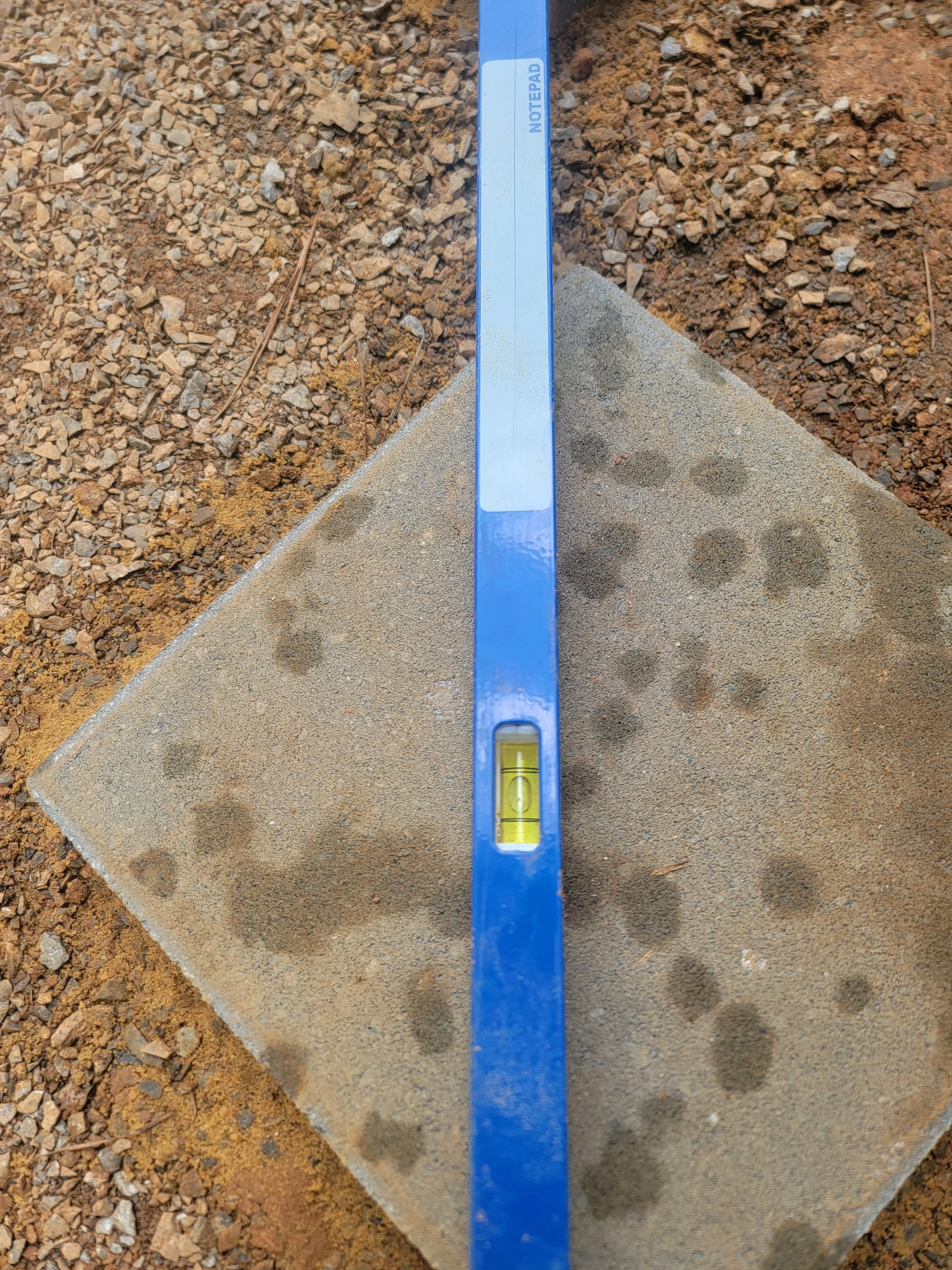 Blue spirit level resting on a concrete paver with dirt and gravel surrounding it.
