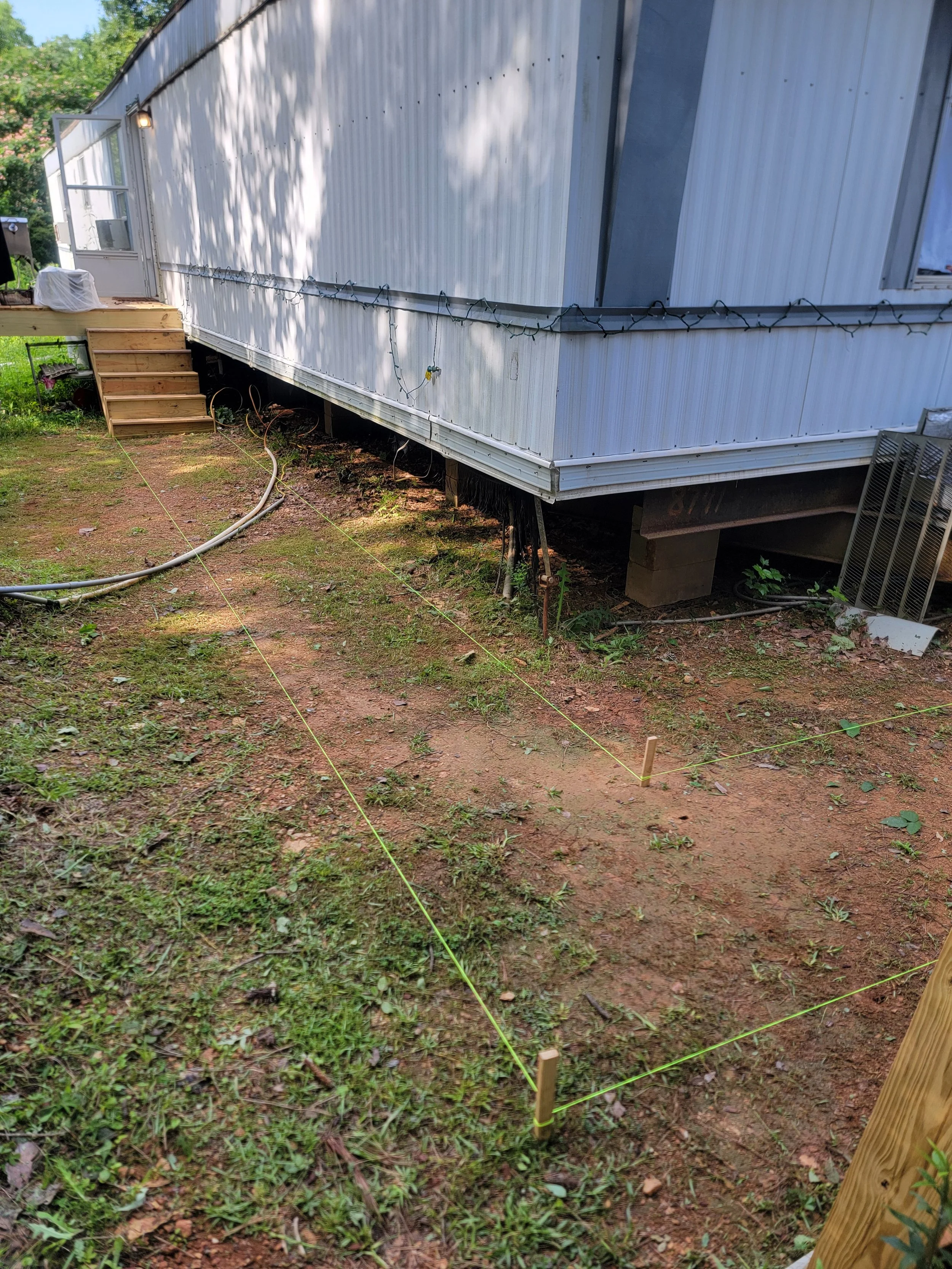 A mobile home with a wooden staircase leading to the entrance, surrounded by green grass and trees. The area is marked off with yellow string tied around wooden stakes, and some electrical cables are on the ground. Shadows of leaves are cast on the side of the mobile home.