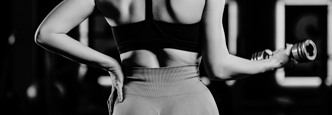 Close-up of a woman's midsection, showing her torso and arms in workout attire, holding a dumbbell at a gym.