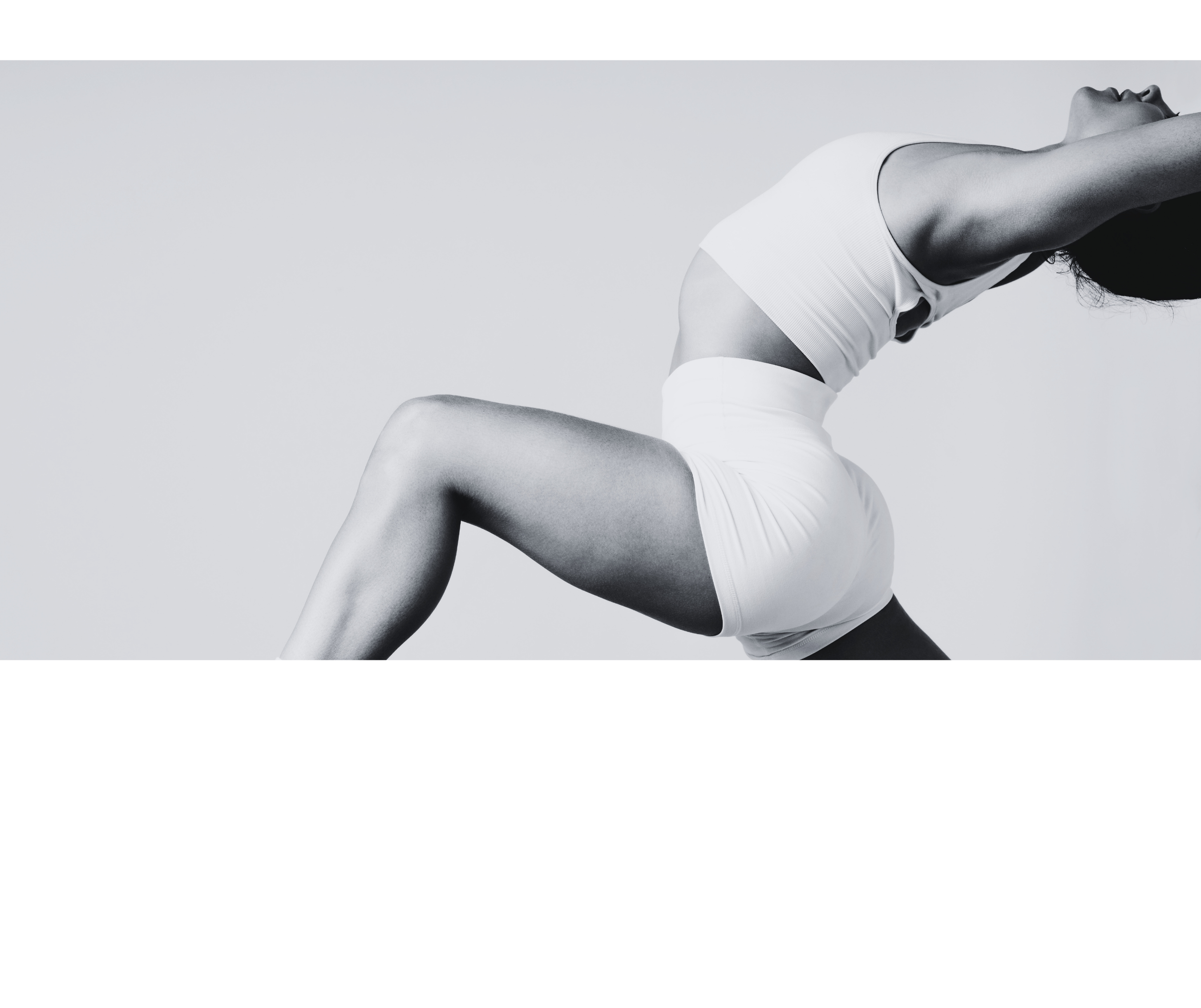 A woman in white athletic wear is performing a yoga pose against a plain light background. She is seated with her knees bent, leaning back with her head tilted upwards, arms stretched behind her, and eyes closed.