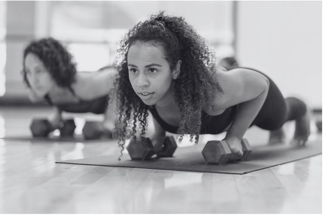 Two women with curly hair doing push-ups with dumbbells on a yoga mat in a gym.