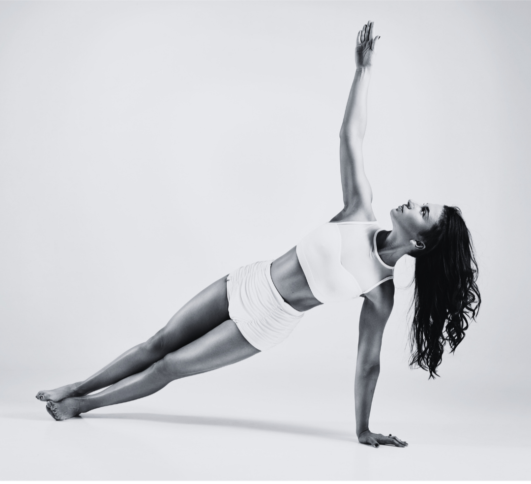 A woman performing a Side Plank Yoga pose on the floor, supporting herself with one arm and raising the other arm upward, looking to her left, wearing a sports bra and shorts, with dark wavy hair and a neutral expression.