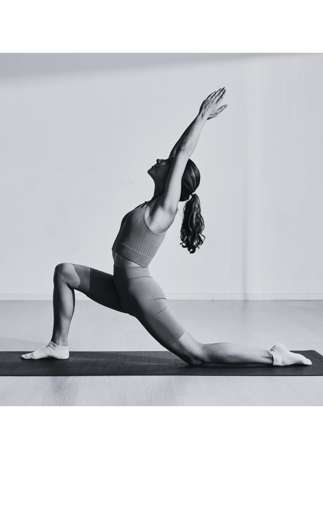 A woman practicing yoga indoors, performing a low lunge with arms extended upward, in front of a plain wall.