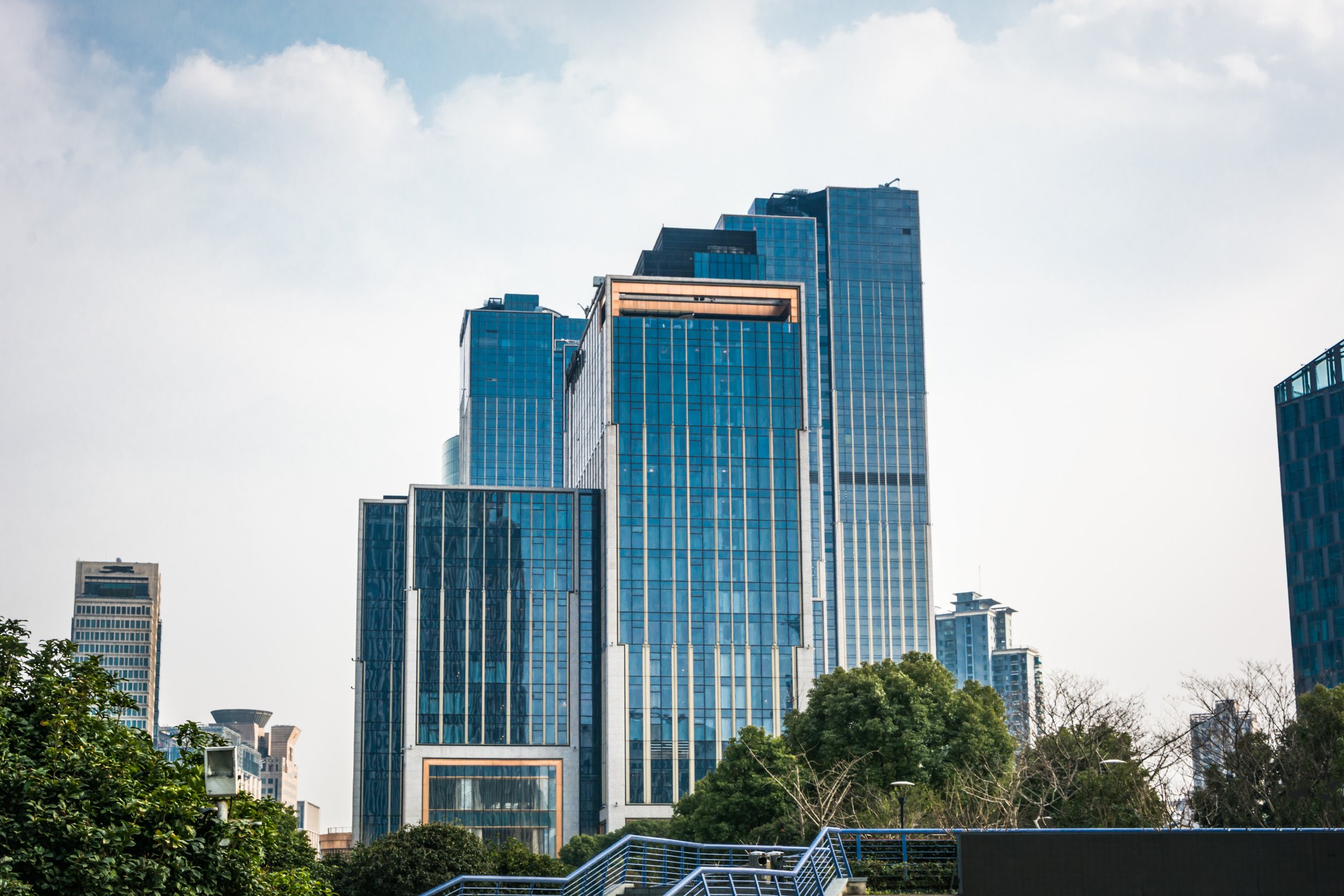 Tall modern glass skyscraper buildings in an urban cityscape with green trees in the foreground and a partly cloudy sky.
