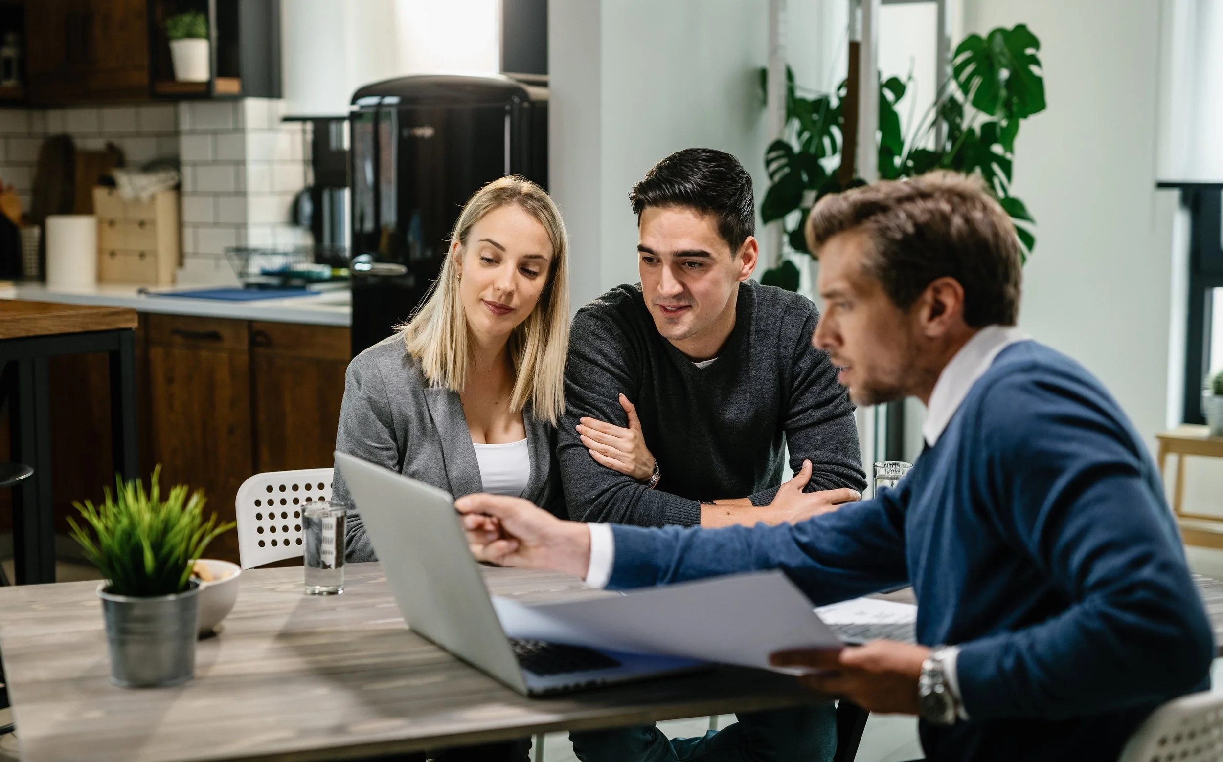 Group of three young professionals sitting at a table in a modern kitchen, focusing on a laptop screen during a discussion or meeting.