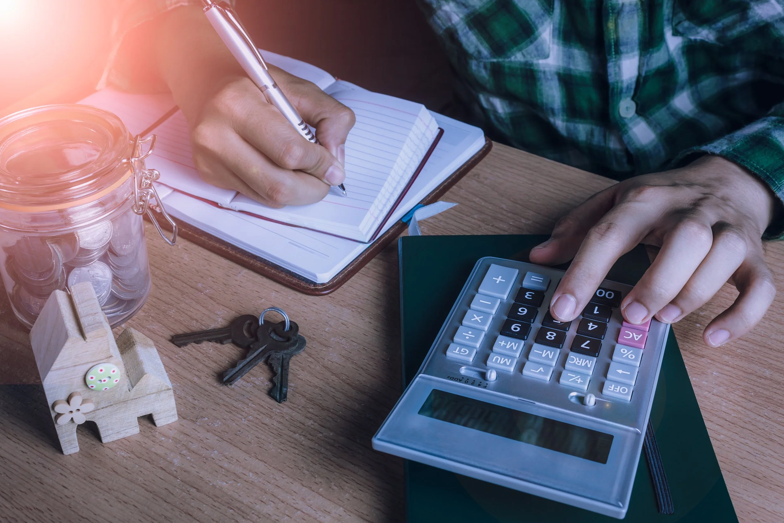 A person wearing a green plaid shirt writing in a notebook with a silver pen. On the wooden desk, there is a jar filled with coins, a set of keys, a small wooden house ornament, a calculator, and some decorative items. The person's hand is pressing buttons on the calculator.