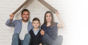 Family of three sitting on the floor under a cardboard box used as a makeshift house, smiling with a white brick wall in the background.