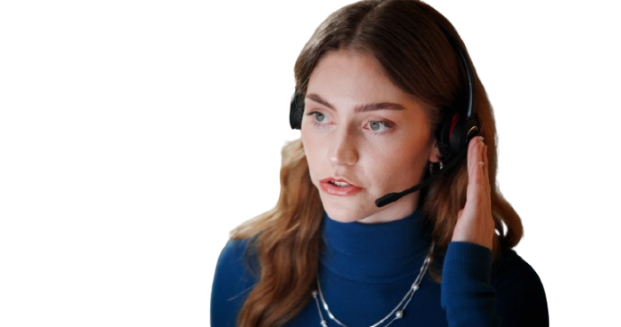 A woman with long brown hair wearing a blue turtleneck and a headset, holding her right hand to the headset as if listening intently.