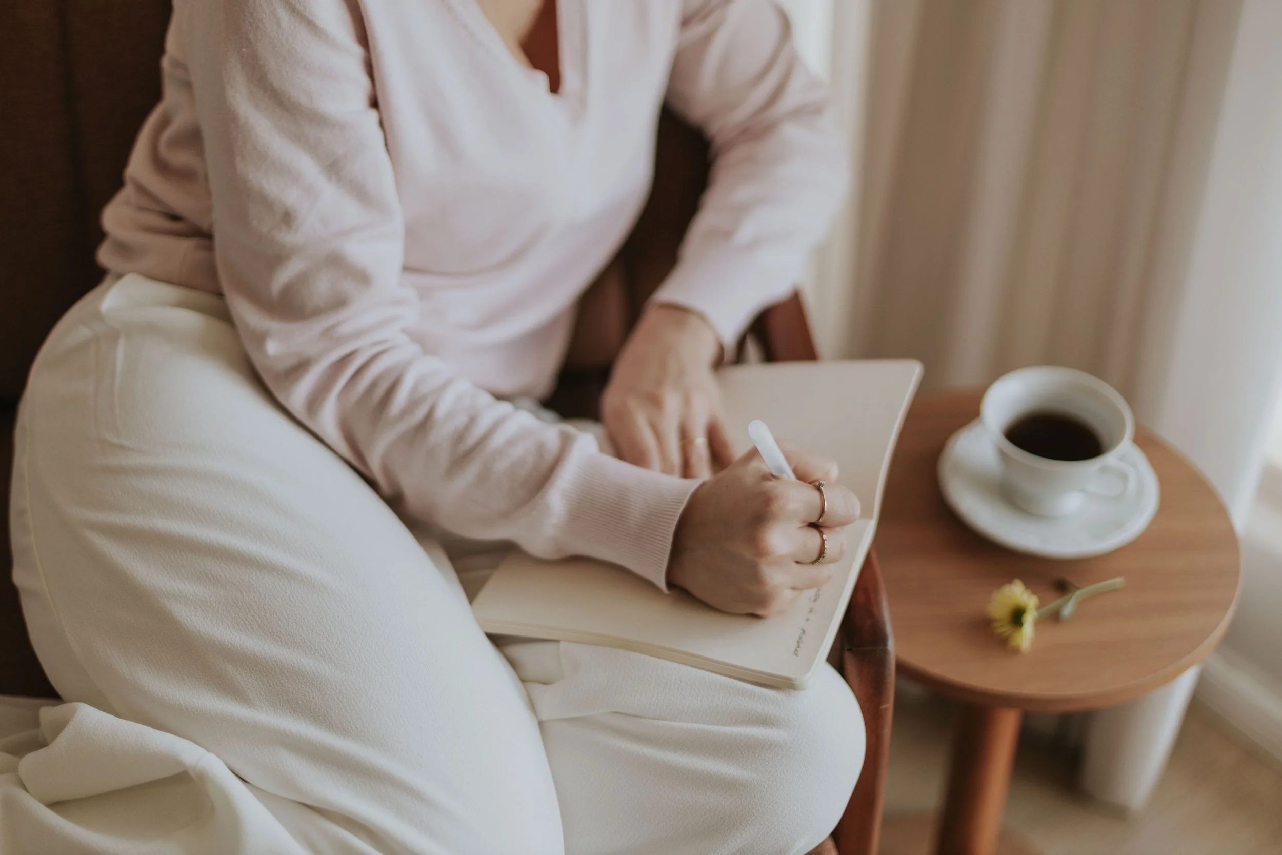 A person wearing a light pink sweater and white pants sitting on a chair, writing in a notebook with a pen. There is a white cup of coffee on a small wooden table next to them, and a small yellow flower on the table.