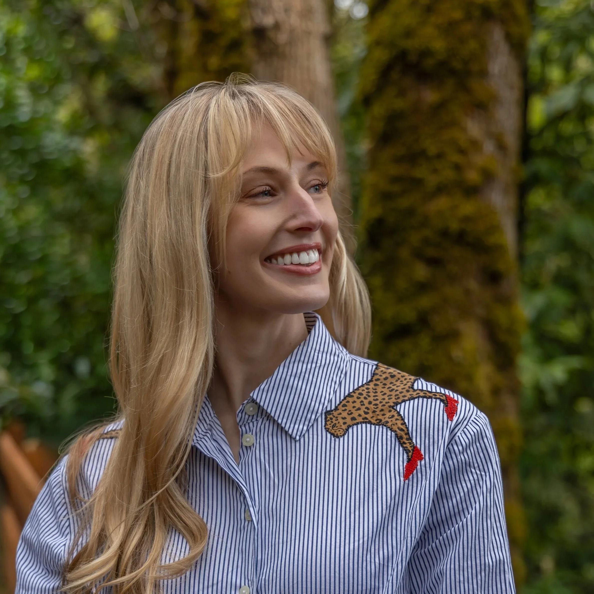A smiling blonde-haired woman outdoors in a wooded area, wearing a blue-and-white striped shirt with a leopard-print detail.