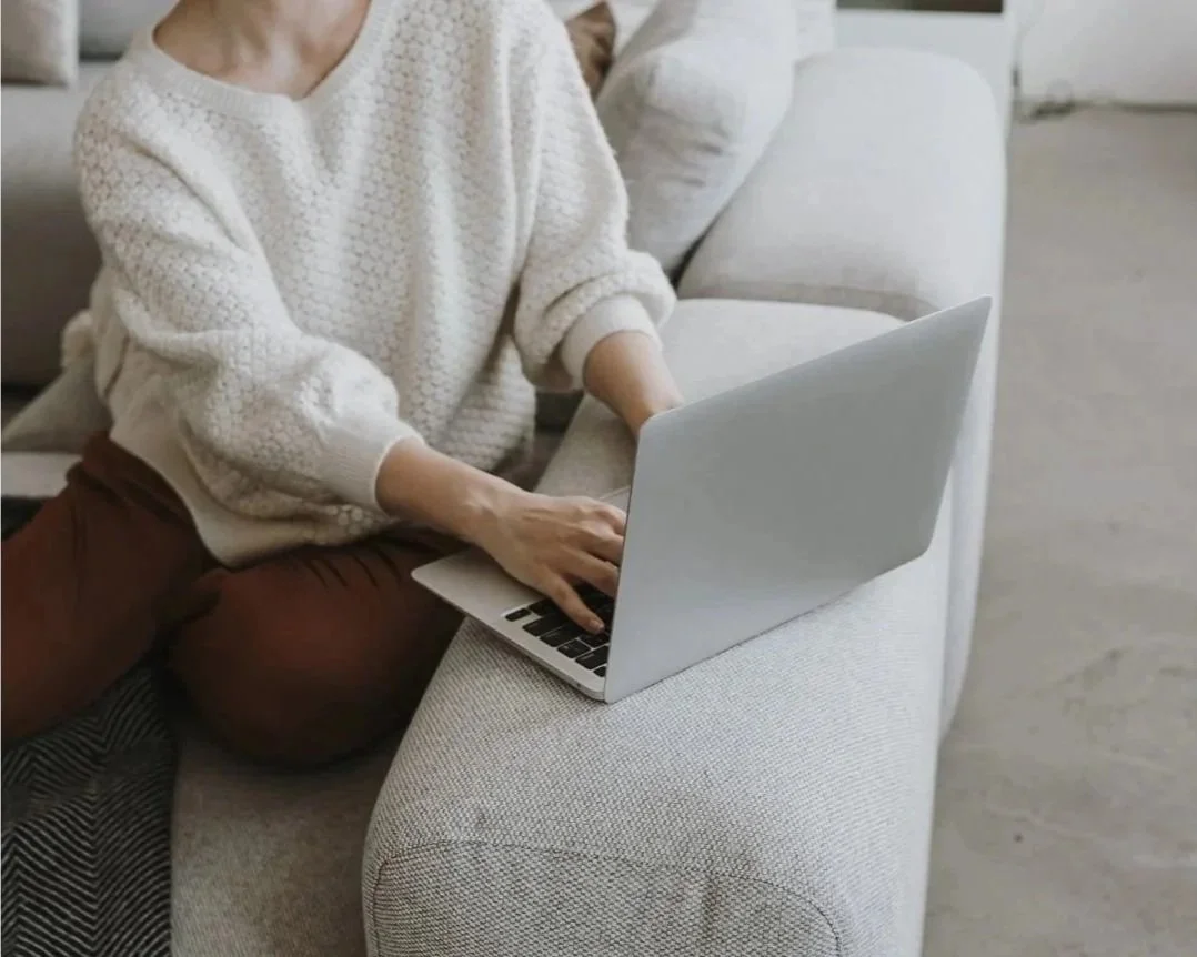 Person sitting on a light-colored sofa using a silver laptop.