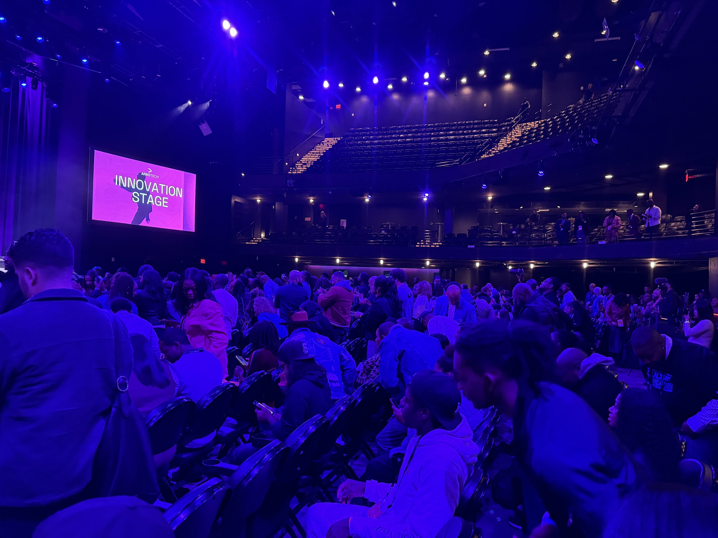 An auditorium stage with a large purple screen that says 'Innovation Stage' and a silhouette of a person, surrounded by an audience seated in rows, illuminated by purple and blue lighting.