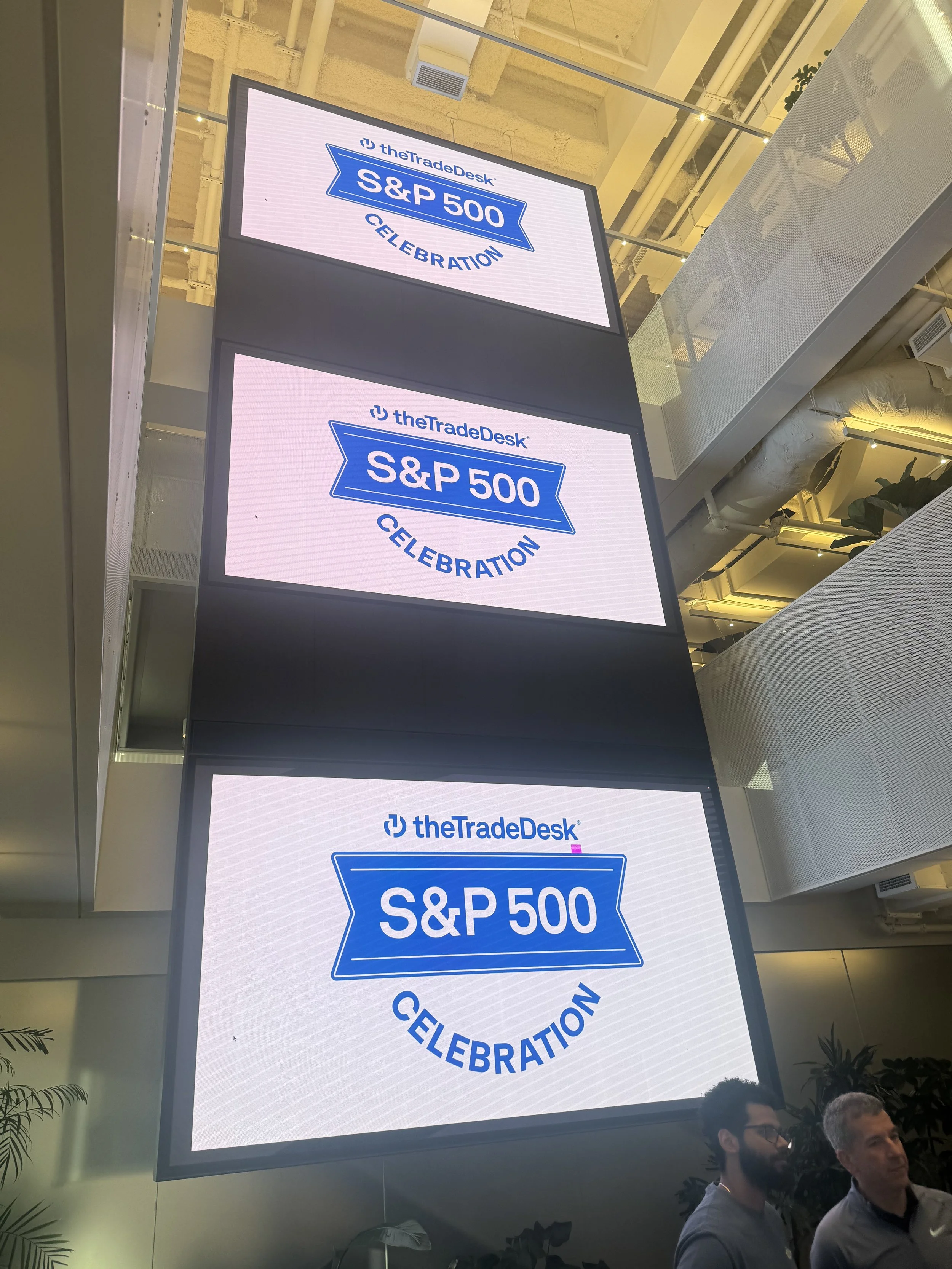 Two large digital screens displaying the logo and text for the S&P 500 celebration event organized by theTradeDesk. The screens are mounted in an indoor venue with ceiling pipes and some people visible at the bottom of the image.