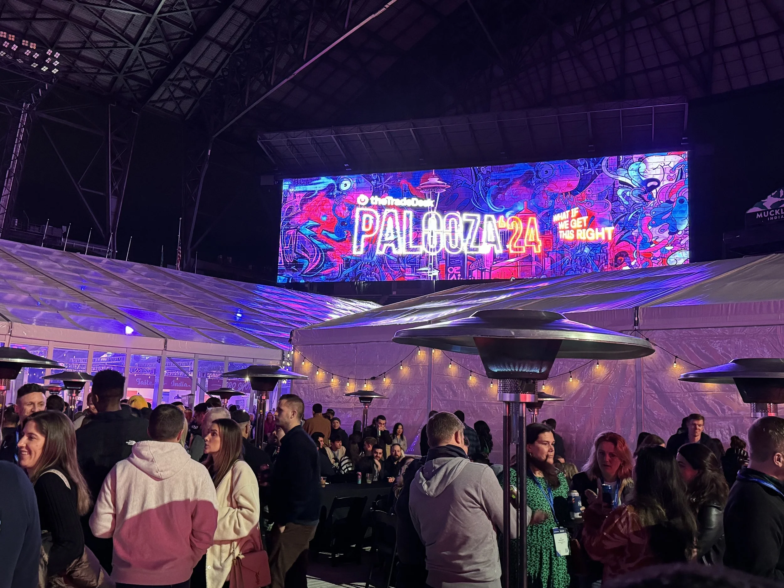 Crowd of people gathered at an indoor music event with a large colorful digital screen displaying the text 'PALOZZIA 24' and 'WHAT IF WE GET THIS RIGHT'.