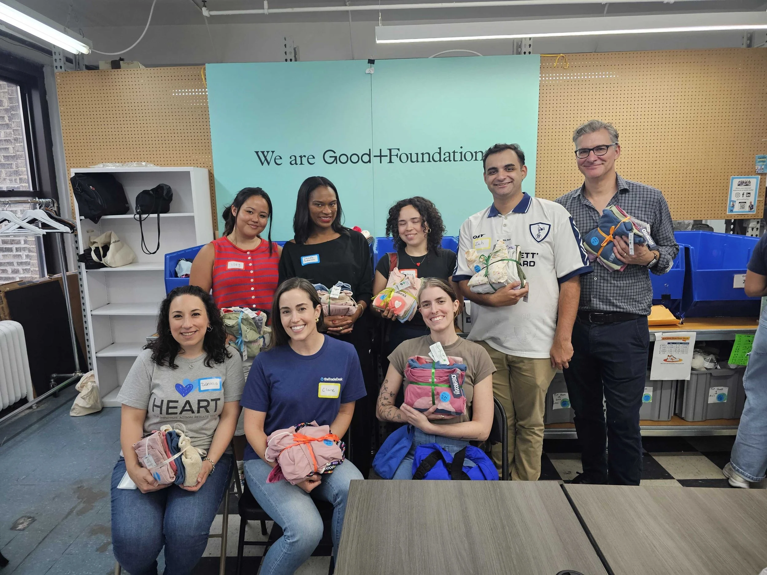 A group of people participating in a charitable event, holding bundles of clothes with tags. They are standing in front of a teal wall with the words "We are Good+Foundation". Some are seated in chairs, and everyone is smiling.