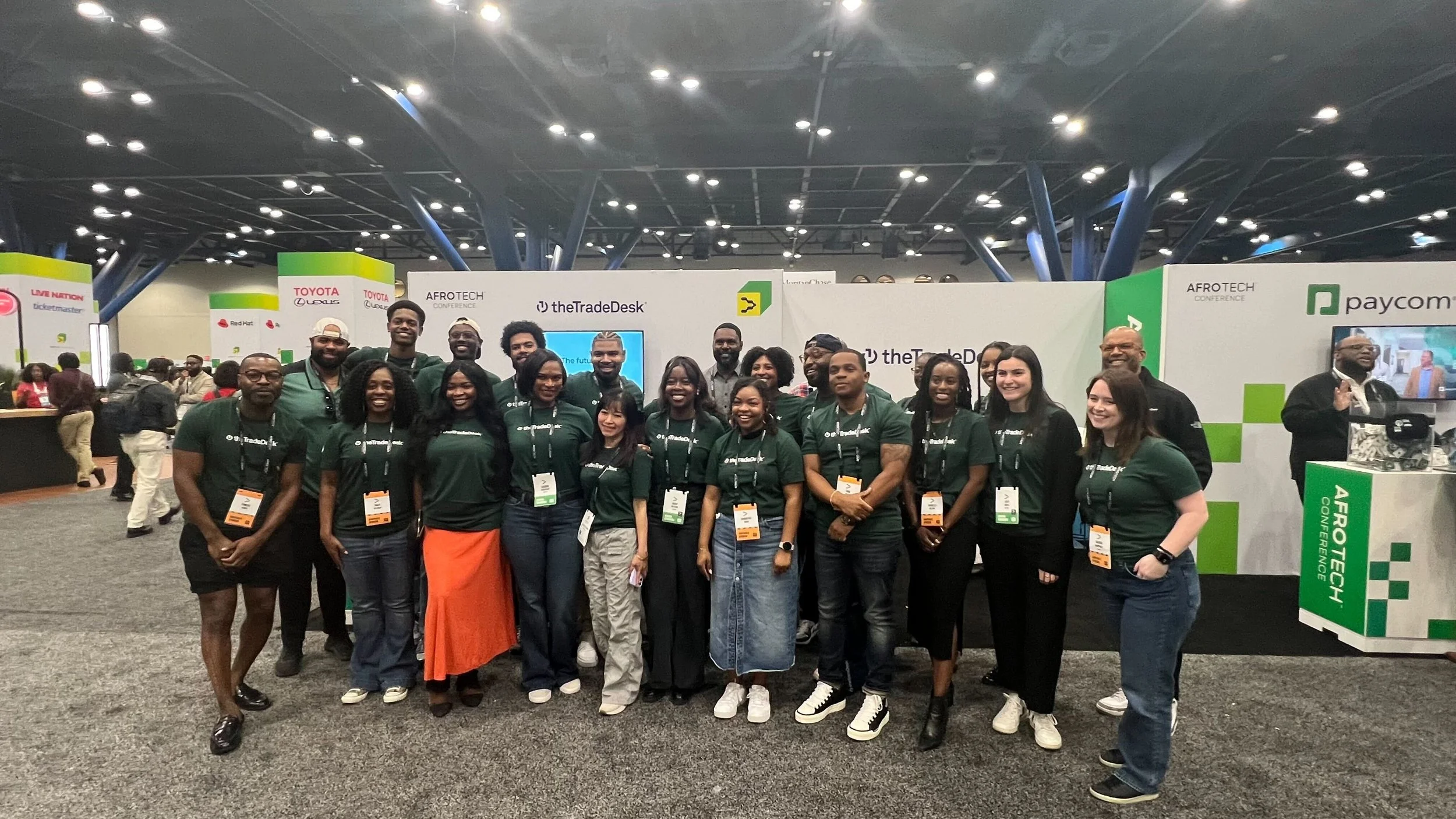 Group of people at a conference standing in front of trade exhibit booths with banners from companies like Paycom and Afrotech, all wearing conference badges and smiling.