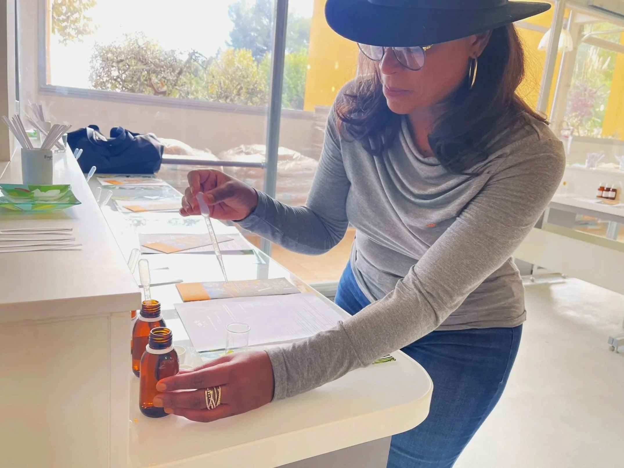 Woman wearing glasses, a wide-brimmed hat, and a gray long-sleeve shirt, handling a dropper bottle in a brightly lit room with large windows and trees outside. Various samples and bottles are on the table.
