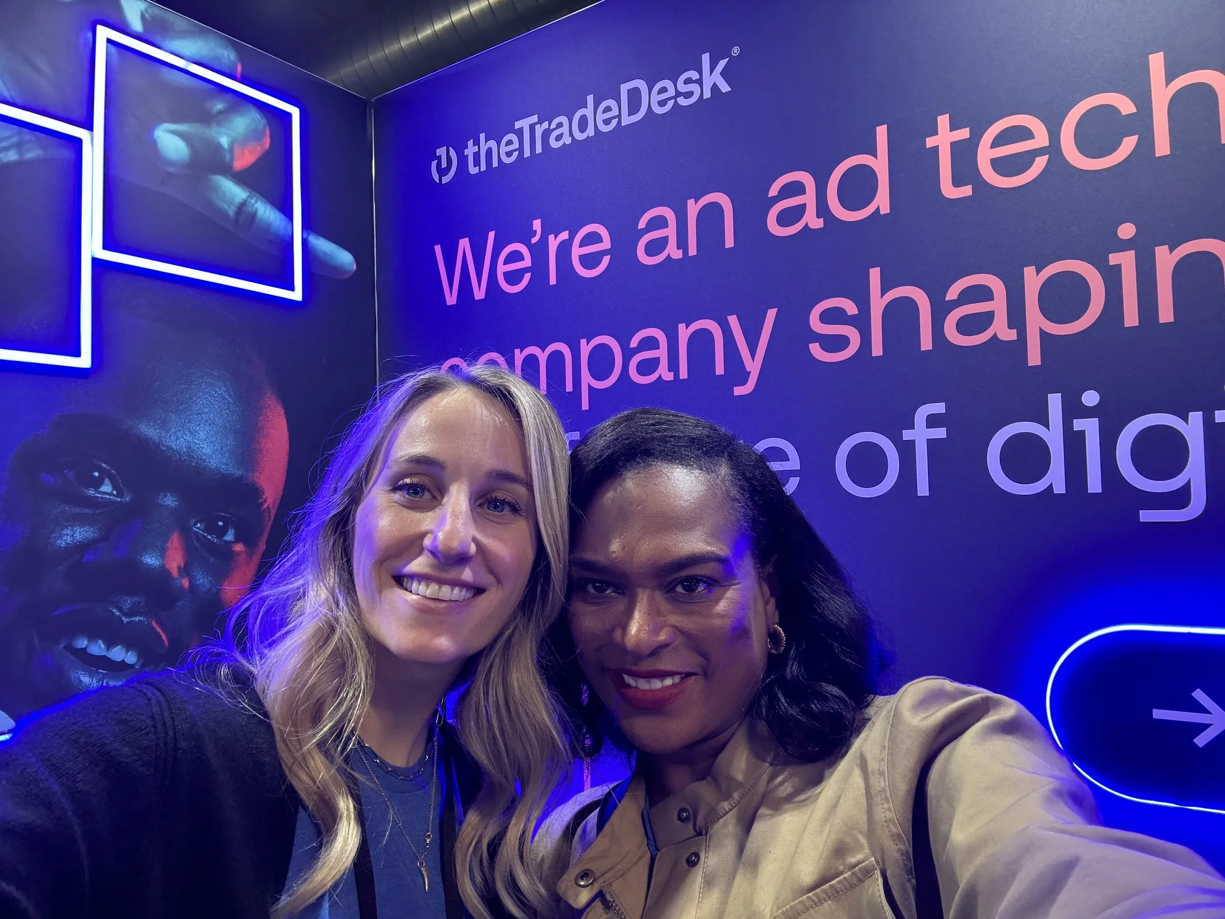 Two women smiling in front of a blue wall with the logo and text for theTradeDesk and the phrase 'We're an ad tech company shaping the future of digital advertising'.