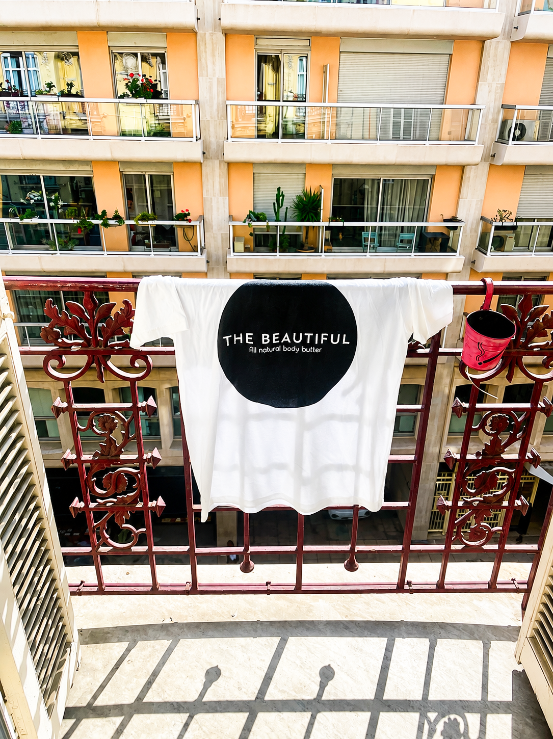 A white t-shirt with a black circle logo that reads 'The Beautiful All Natural Body Butter' is hanging on a balcony railing, with a pink bucket attached. In the background, there is an apartment building with potted plants on the balconies.