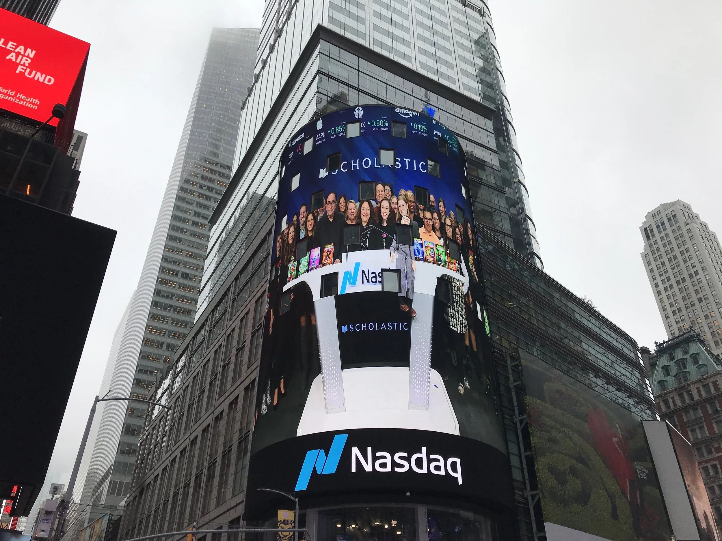 Tall building in Times Square with digital billboards, including Nasdaq and Scholastic advertisements, and a cloudy sky in the background.