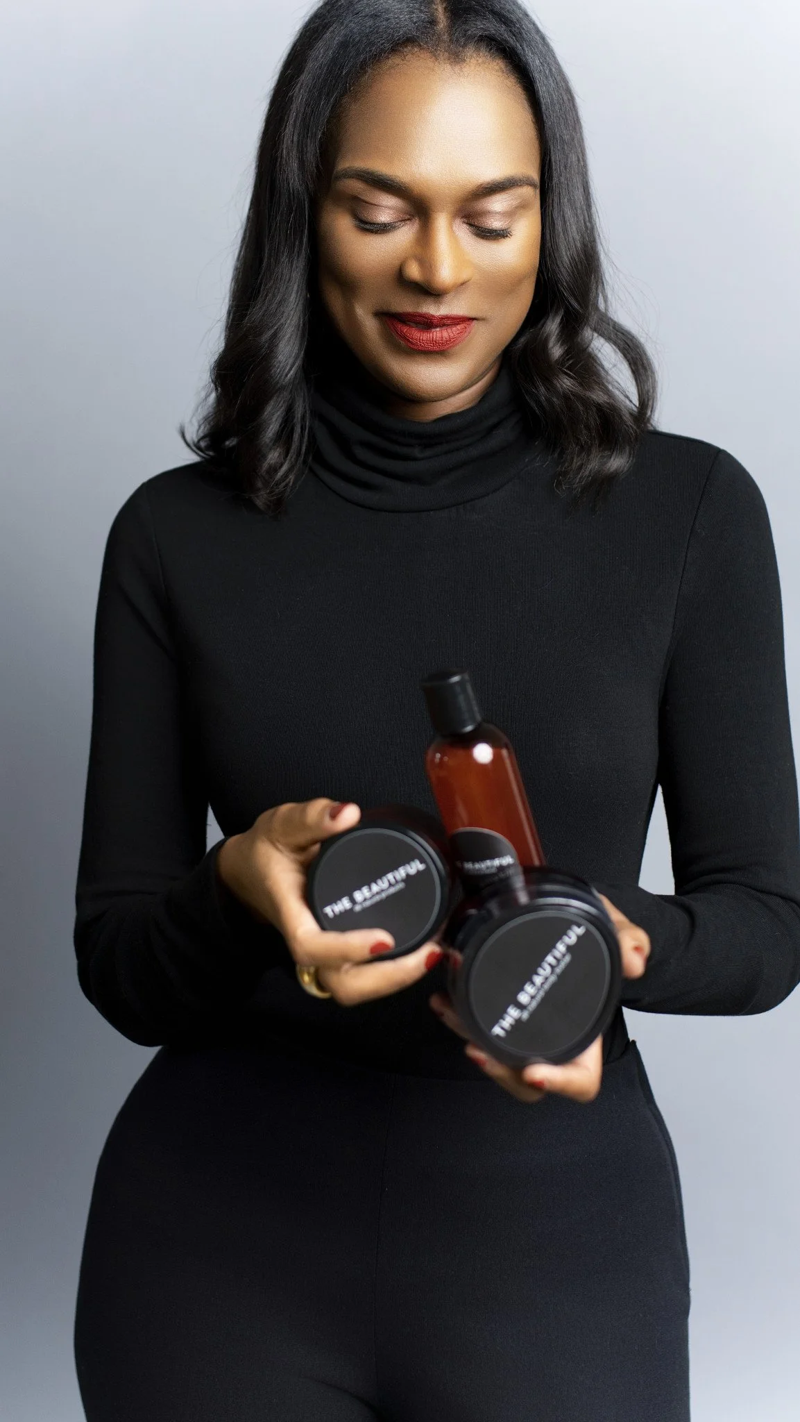 A woman with dark hair and red lipstick wearing a black turtleneck holding skincare or beauty product containers labeled 'THE BEAUTY' against a gray background.